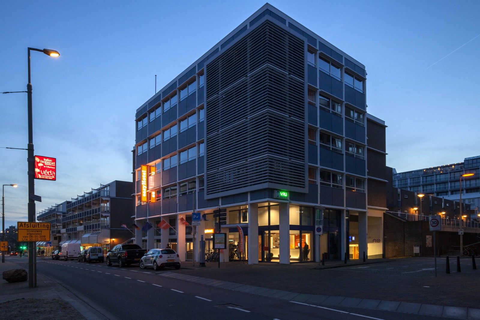 Exterior view of the modern easyHotel The Hague Scheveningen Beach at dusk, featuring illuminated orange easyHotel signs along a city street.