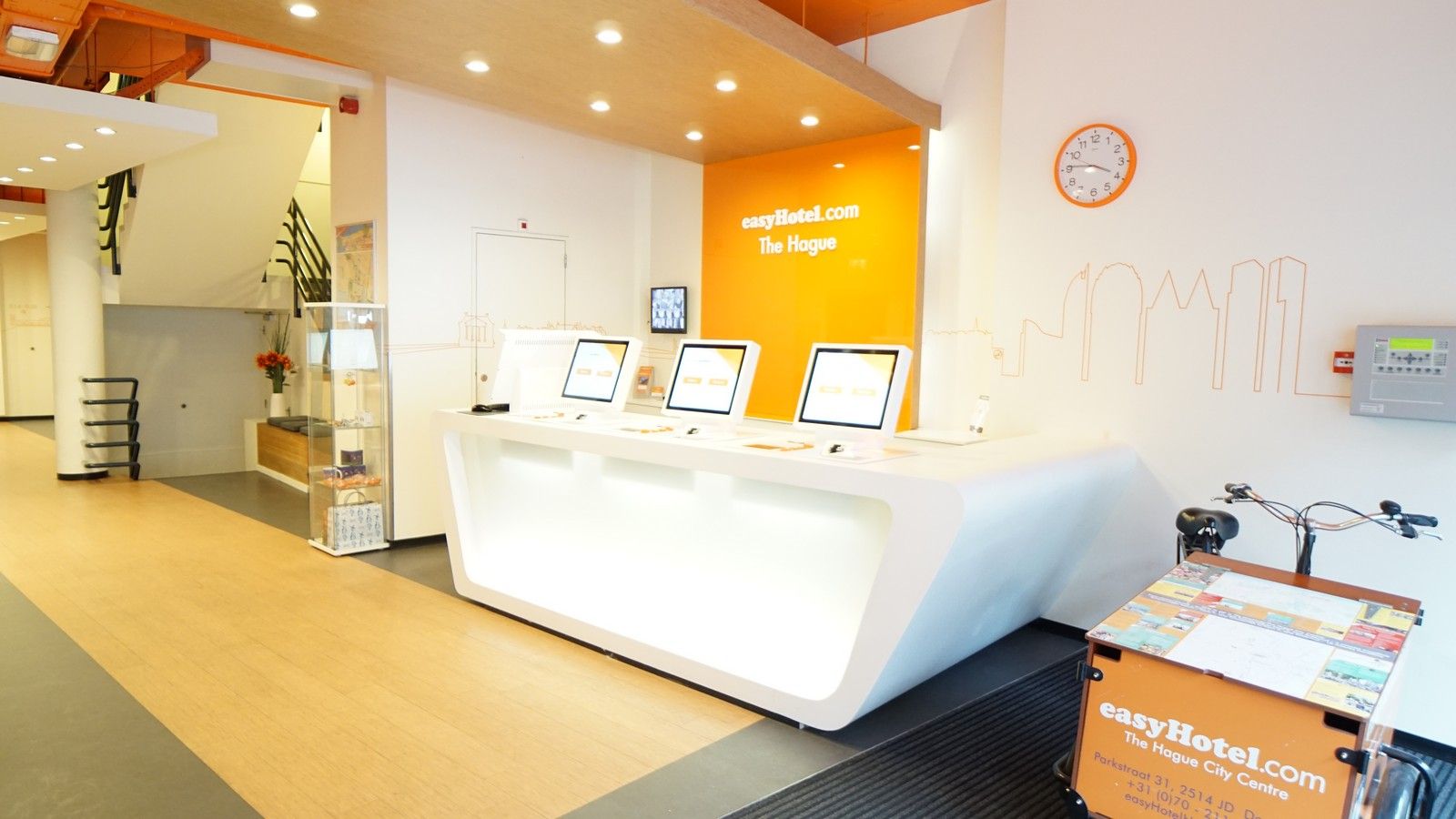 Bright, modern easyHotel lobby in The Hague featuring white self-service check-in kiosks, vibrant orange branding, and a cityscape wall graphic.