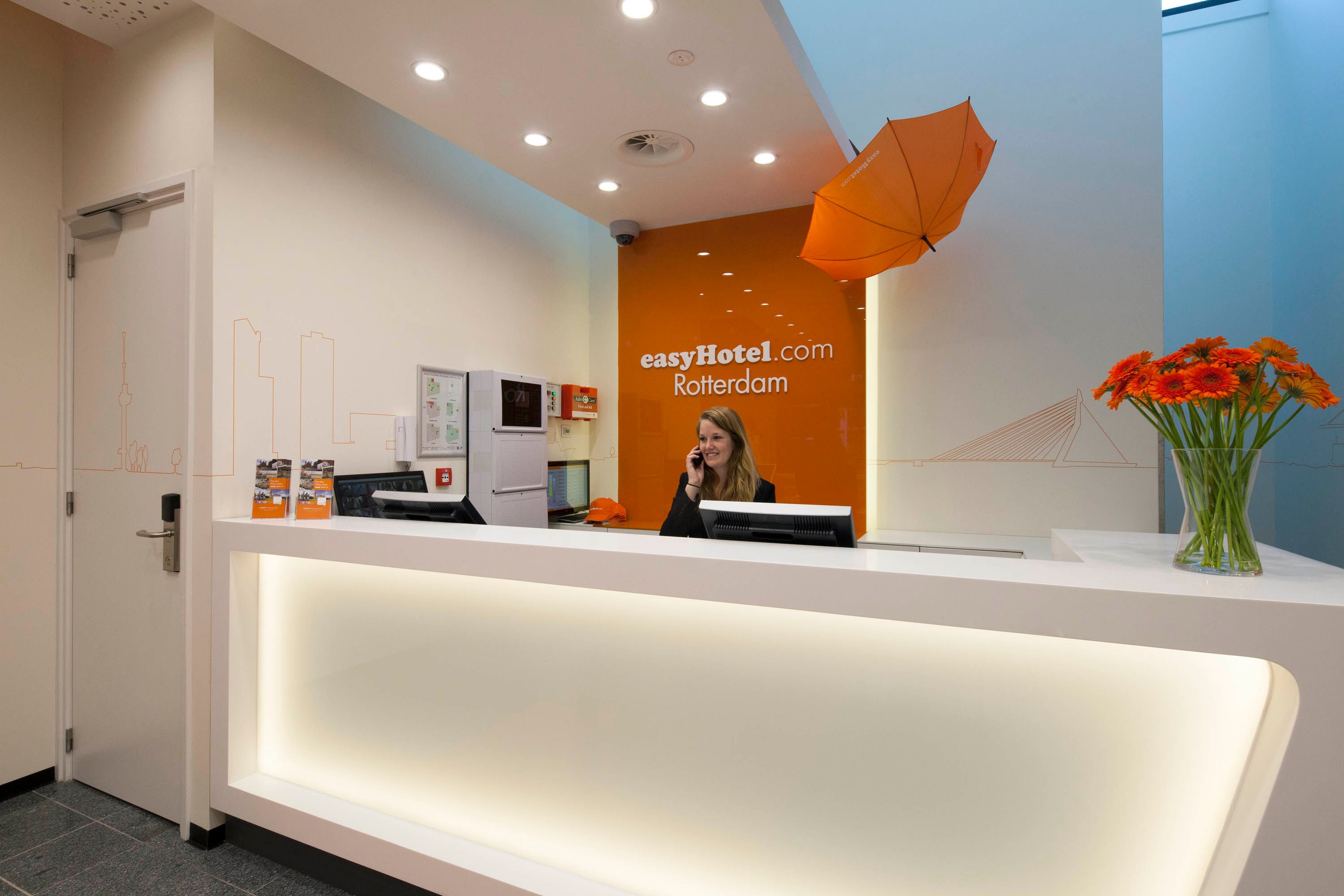 Brightly lit easyHotel Rotterdam reception desk featuring a smiling female hotel employee on the phone, an orange wall with the easyHotel.com Rotterdam logo, and a decorative orange umbrella.