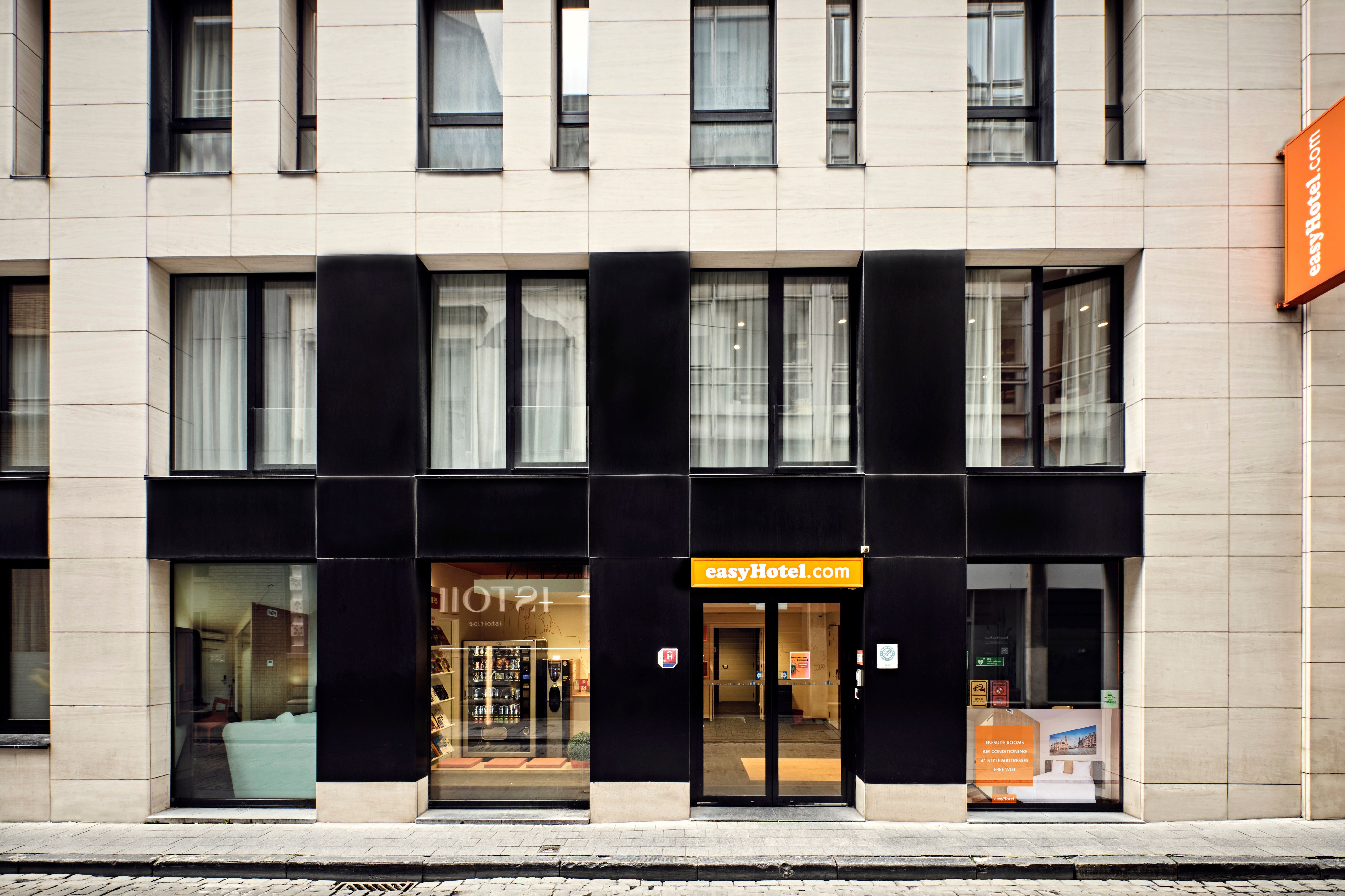 Exterior of the easyHotel Brussels City Centre, showcasing its modern facade with light-colored stone and dark black sections, large windows, and a prominent orange easyHotel.com sign above the entrance.