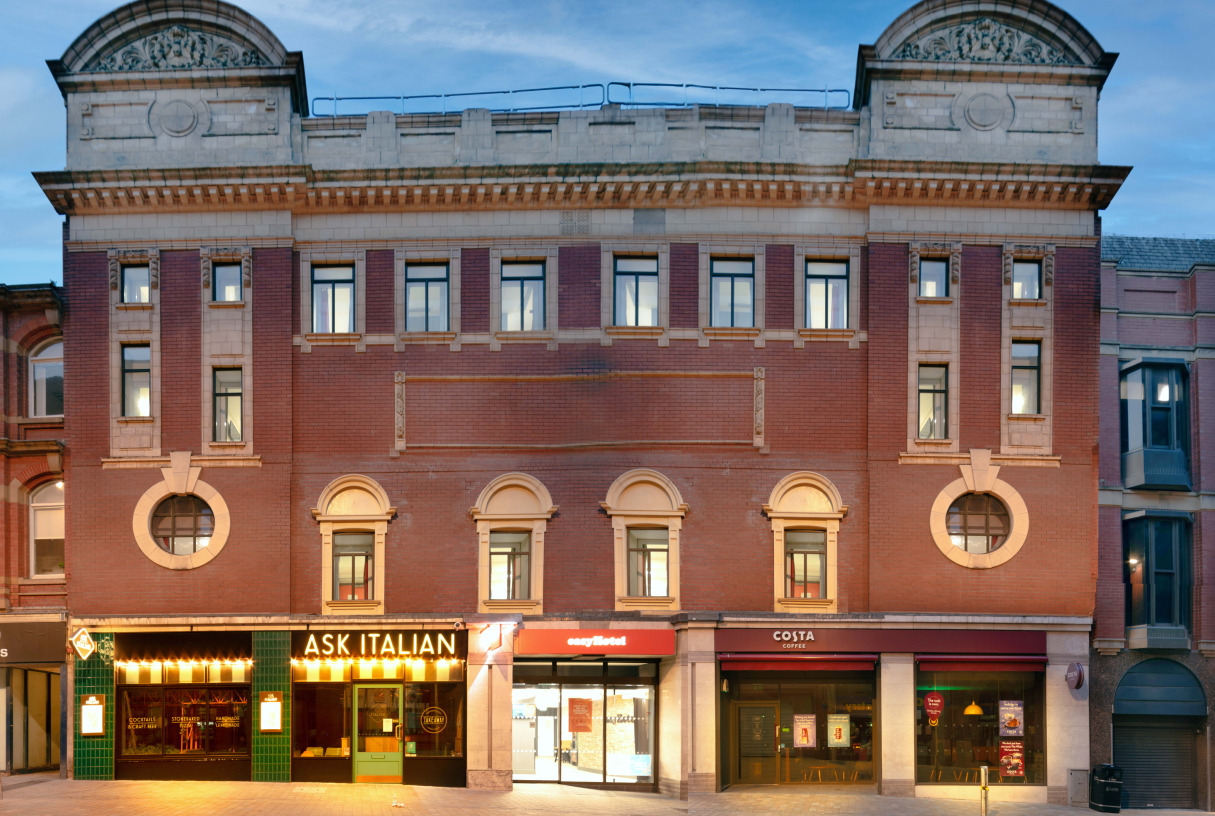 Facade of easyHotel Leeds, a prominent red brick building with ornate light stone architectural details and multiple windows. The ground floor features entrances for easyHotel, ASK ITALIAN, and Costa Coffee.