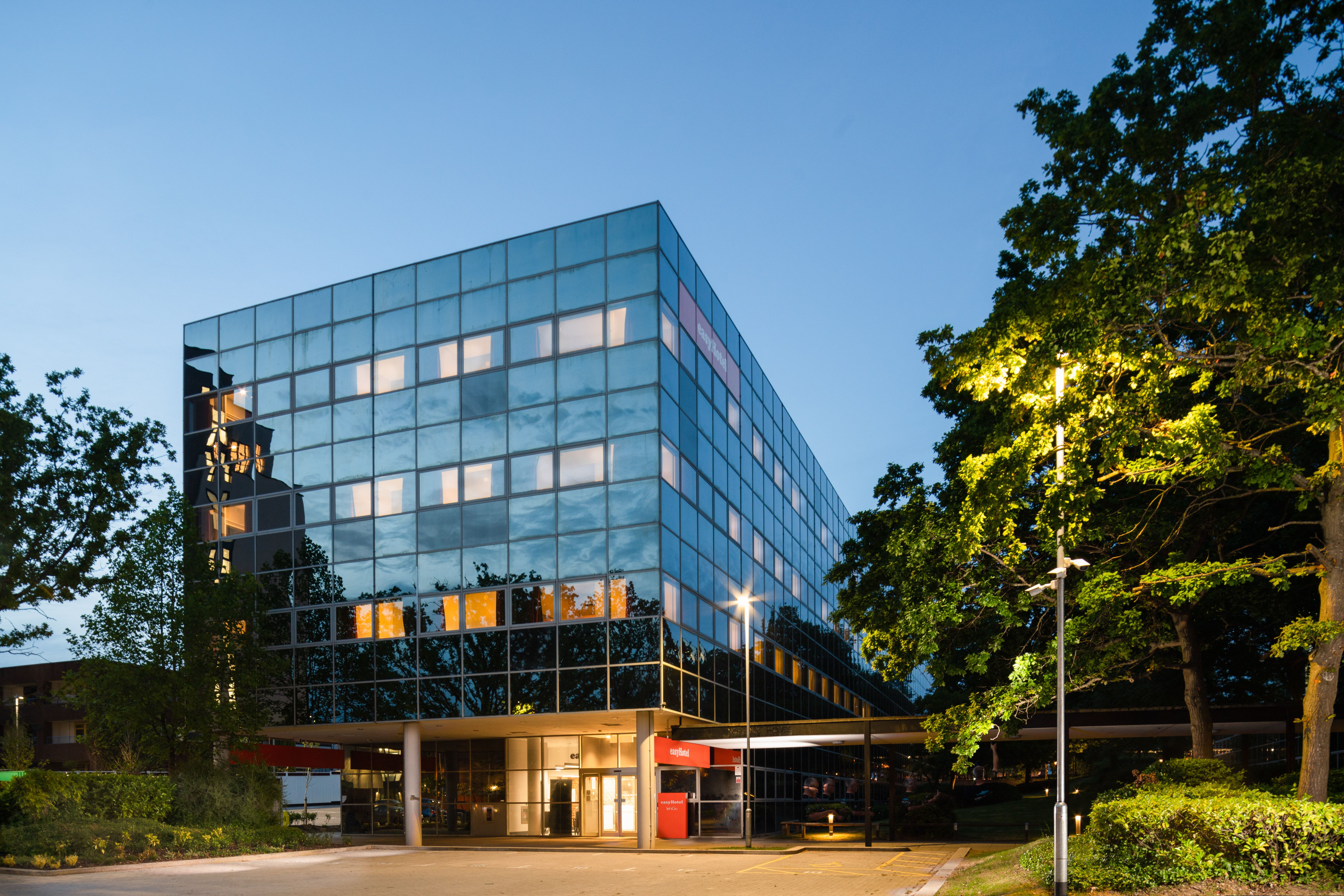 easyHotel Milton Keynes exterior: Modern glass-fronted hotel building with illuminated windows at dusk, surrounded by green trees.