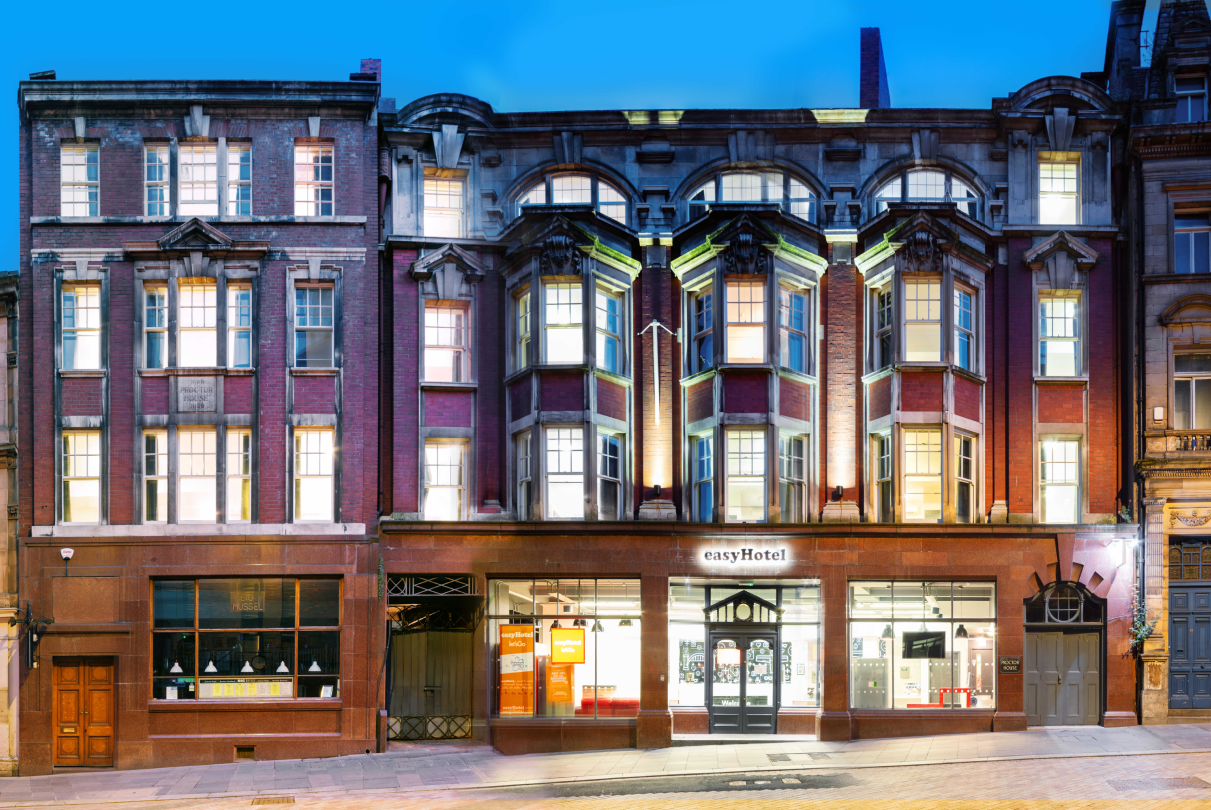 Exterior of easyHotel Newcastle at dusk, showcasing its traditional red brick facade with ornate bay windows and a brightly lit ground floor entrance.