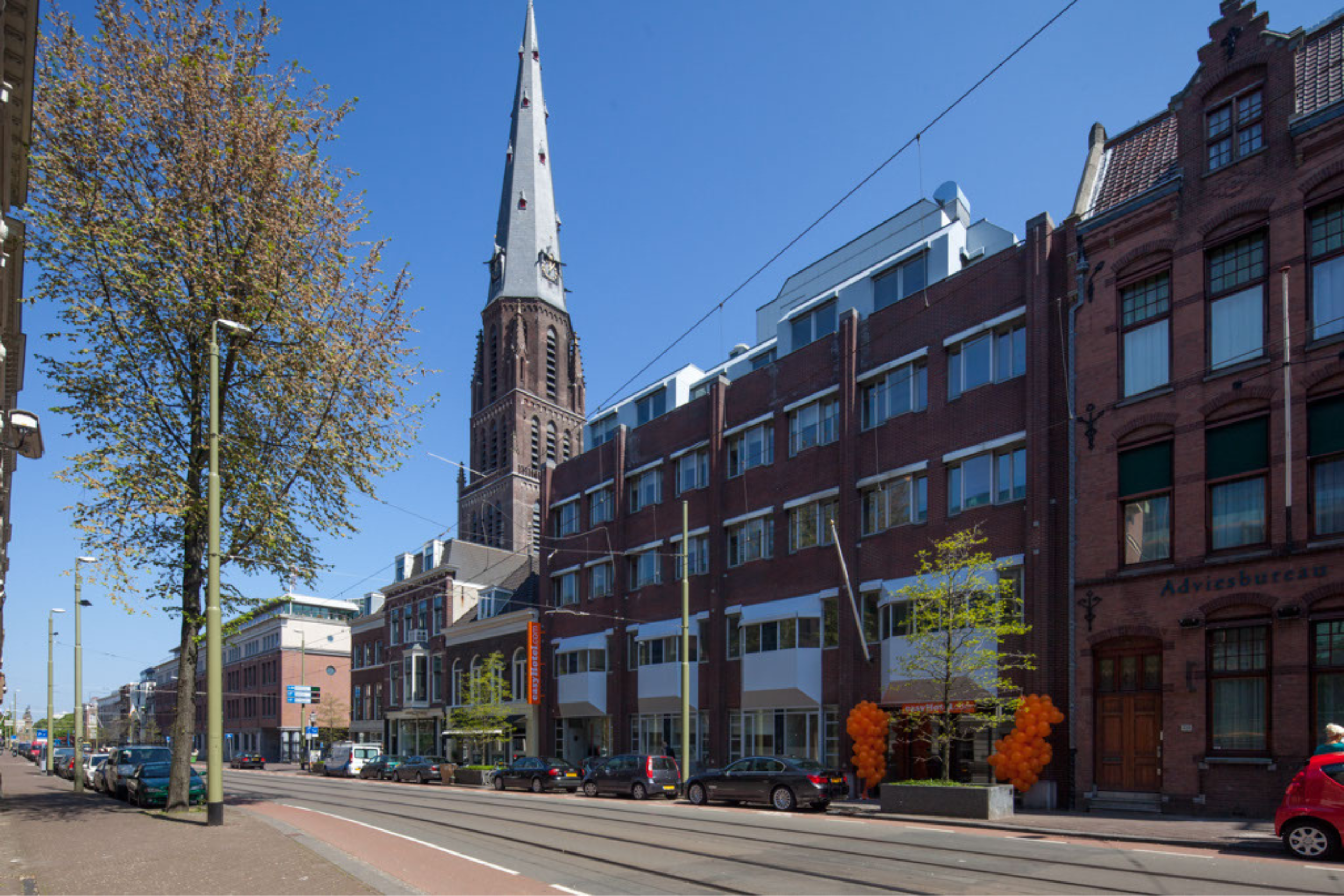 Exterior view of easyHotel The Hague building on a sunny city street with tram lines, cars, and the historic Grote Kerk steeple prominent in the background.