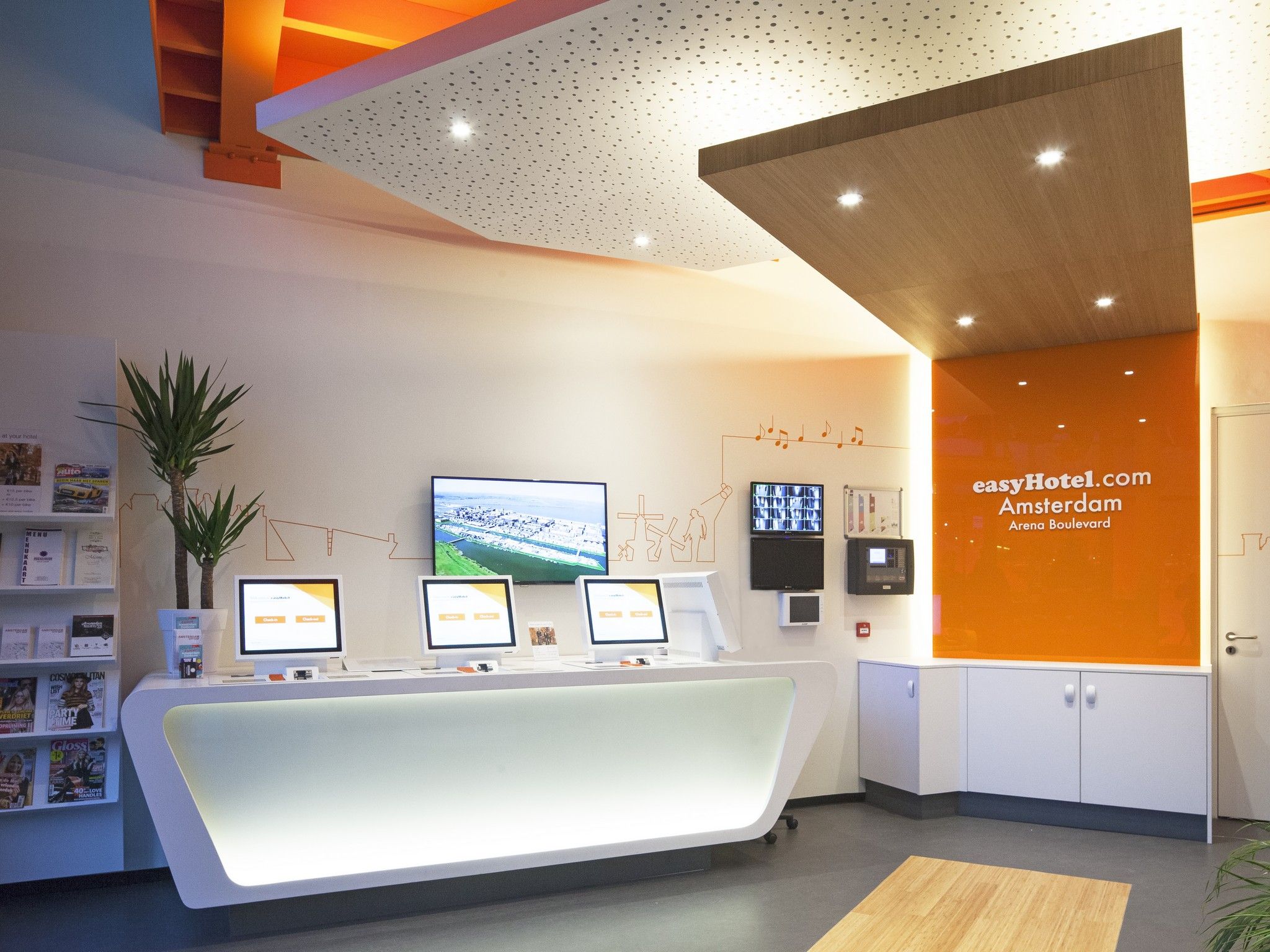 Modern easyHotel Amsterdam Arena Boulevard reception area featuring a white, illuminated desk with computers, and a prominent orange wall displaying the hotel's branding.