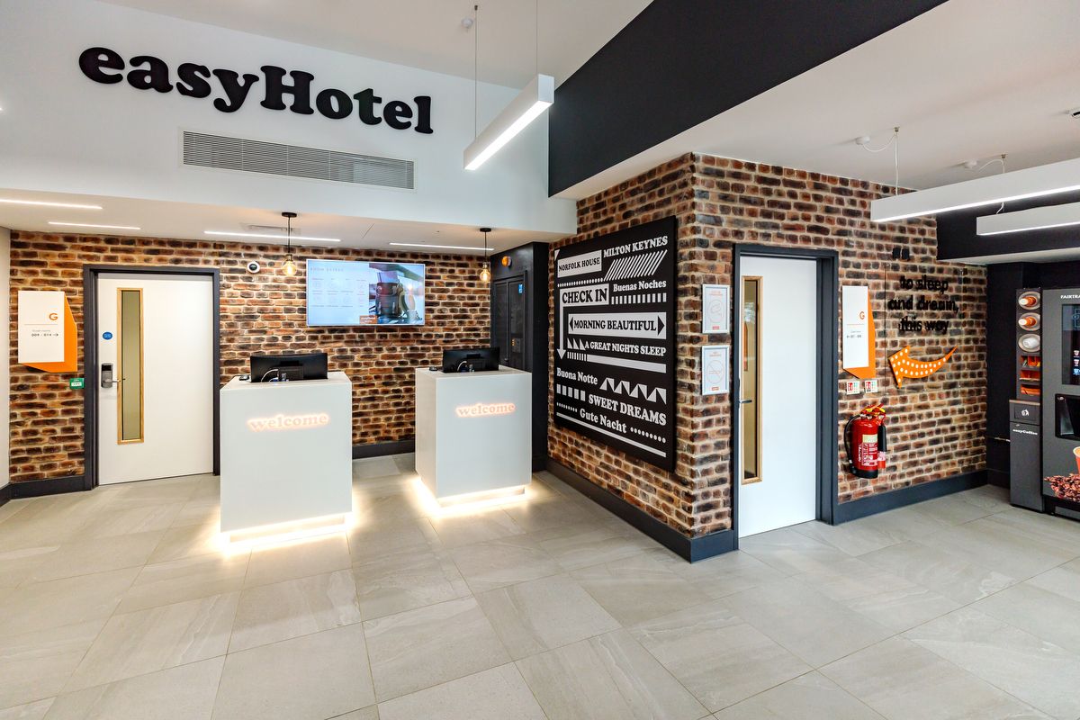 Bright, modern easyHotel Milton Keynes reception area featuring exposed brick walls, light grey tiled flooring, and two self-service check-in kiosks with illuminated 'welcome' signs.