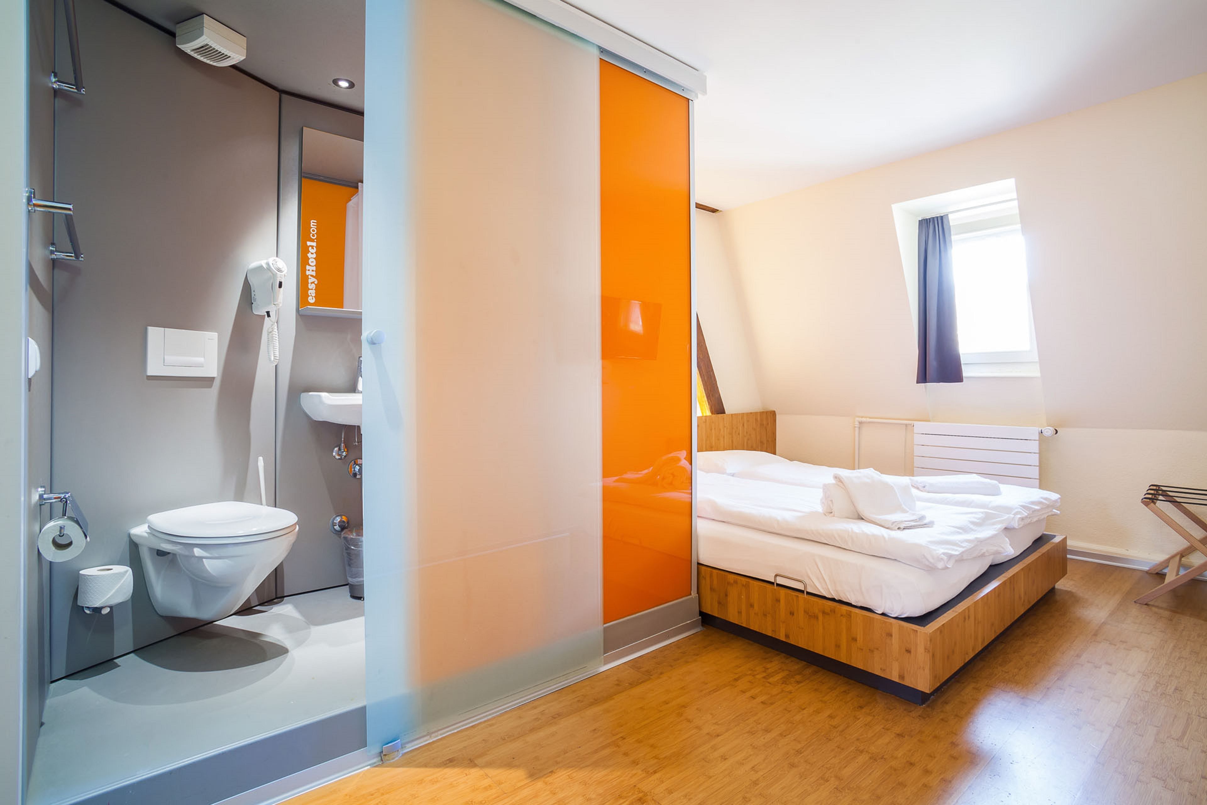 Modern easyHotel room with a double bed, white bedding, and a compact bathroom area featuring a toilet and sink, separated by a frosted glass and orange sliding door. The room has wooden flooring and a window.