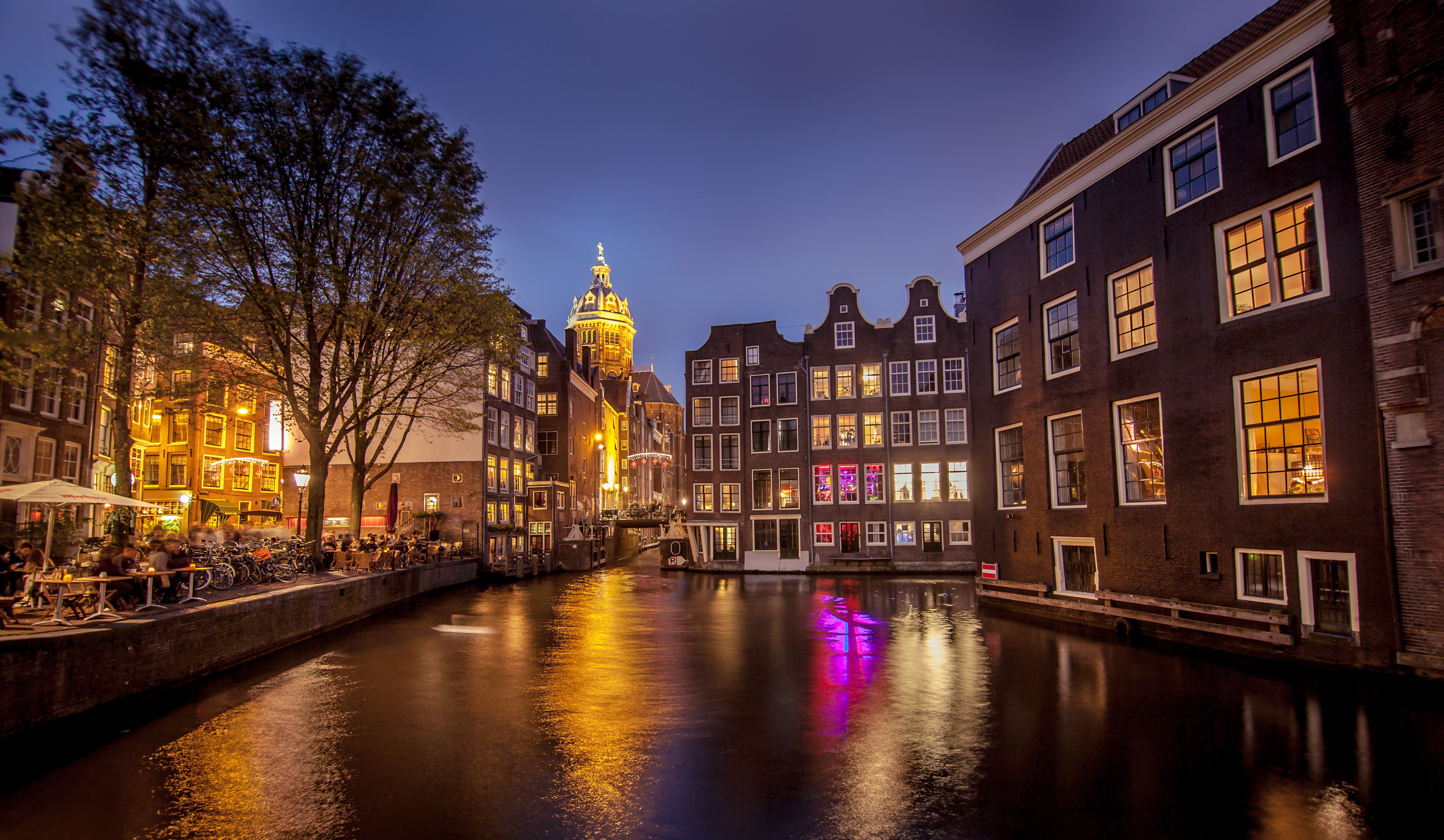 Night view of an Amsterdam canal, featuring illuminated historic canal houses with vibrant bars and restaurants, reflections shimmering on the water, and the dome of the Basilica of Saint Nicholas in the background.