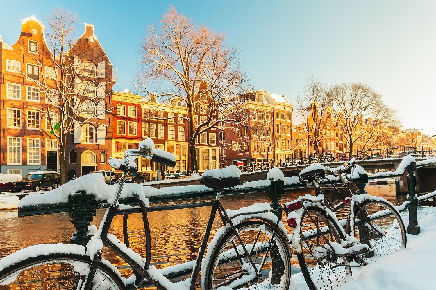 Snow-covered bicycles parked along an Amsterdam canal with historic gabled houses and bare trees under a clear winter sky.