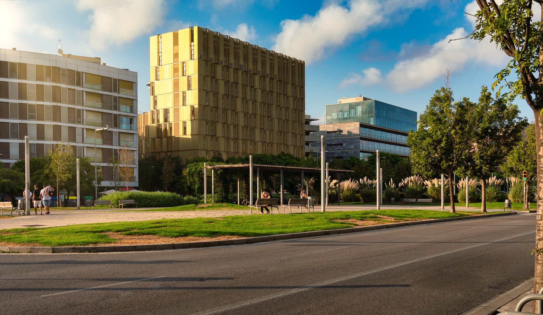 A vibrant Barcelona urban scene showcasing a golden-yellow modern building, contemporary architecture, a lively green park with people, and a street on a sunny day.