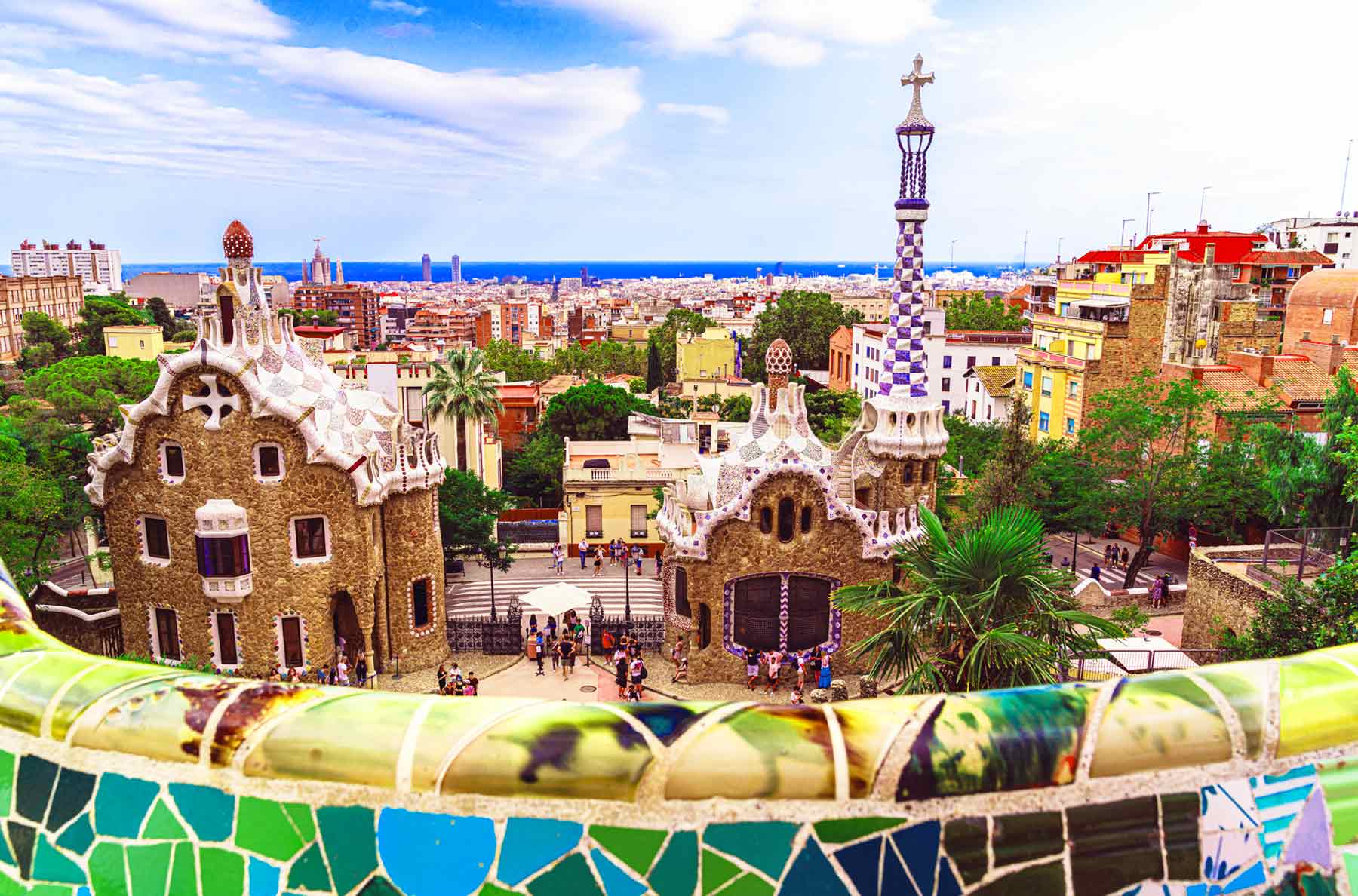 Panoramic view of Antoni Gaudí's Park Güell in Barcelona, featuring the whimsical Porter's Lodge and mosaic-tiled railing, with the city and Mediterranean Sea in the background.