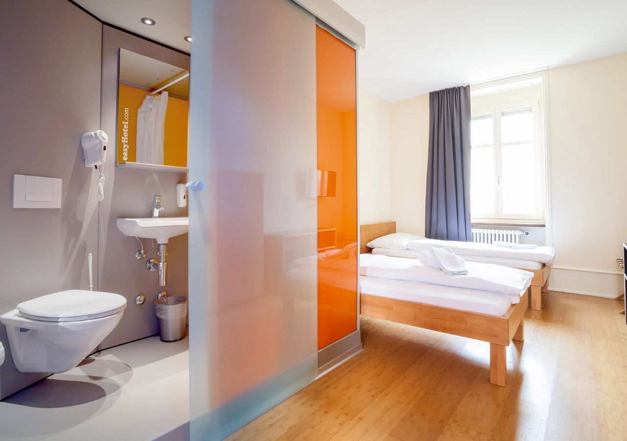 An easyHotel room in Basel with two single beds, white bedding, and a modern compact bathroom featuring a toilet, sink, and orange-accented frosted sliding door.