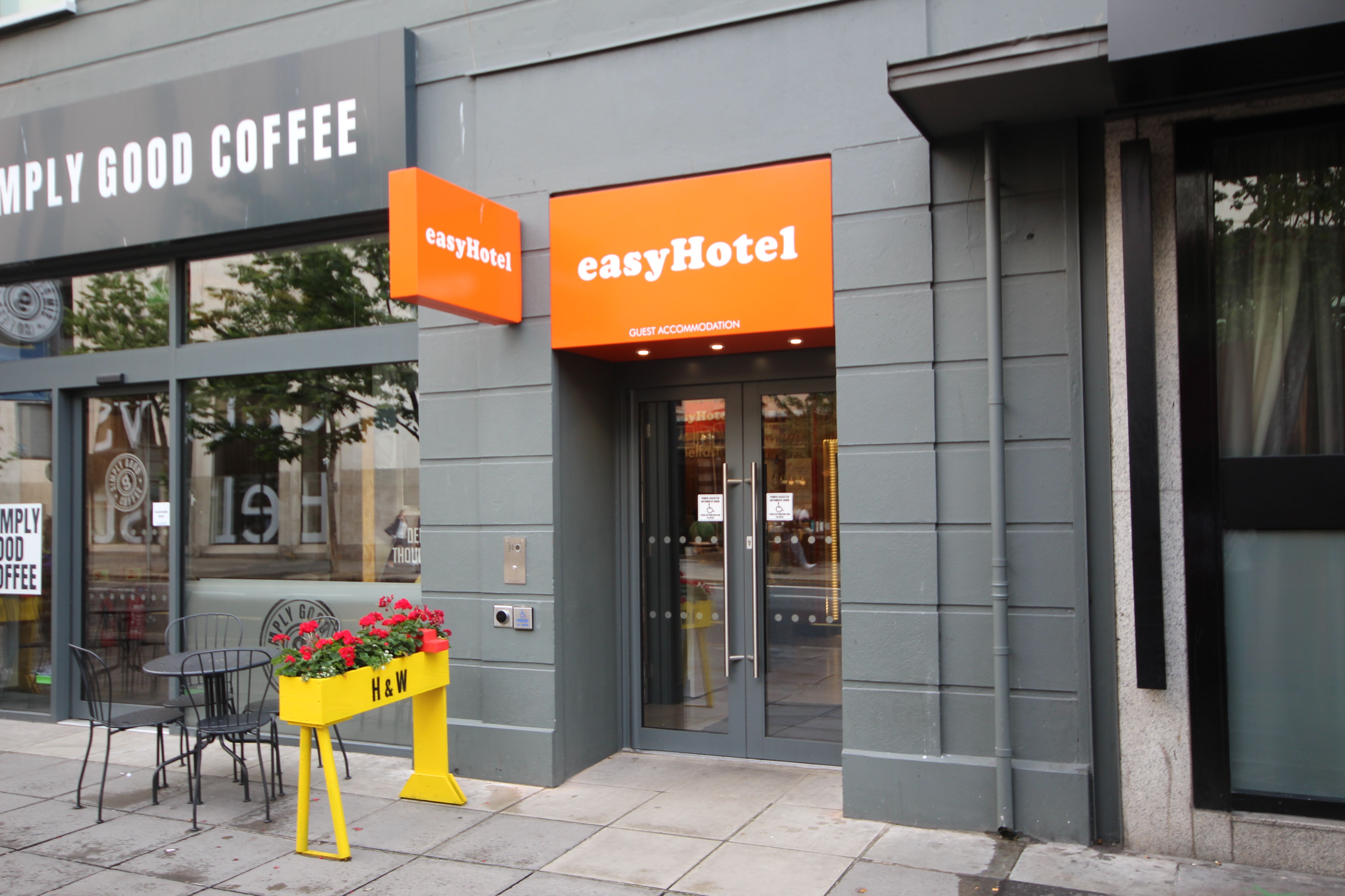 Exterior view of the easyHotel entrance in Belfast. Bright orange easyHotel signs are visible above the glass double doors. To the left, a coffee shop with 'Simply Good Coffee' branding has outdoor seating with a black metal table and chairs. A yellow pla
