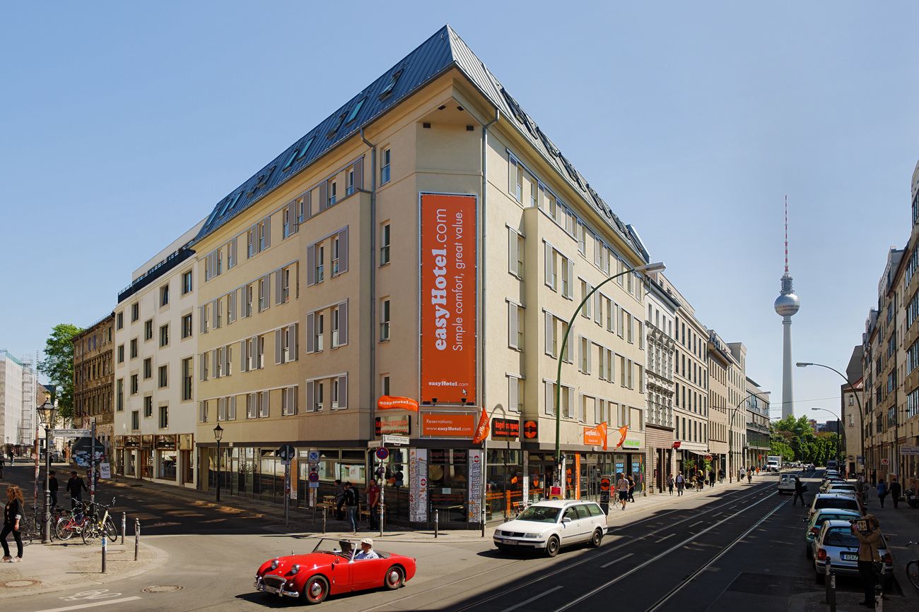 Exterior view of the easyHotel in Berlin, located on a street corner, with a red classic car driving by and the Berlin TV Tower visible in the background.