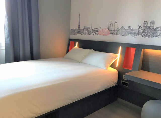 EasyHotel room in Paris featuring a double bed with white bedding, integrated yellow LED headboard lighting, and a distinct Parisian skyline mural on the wall.