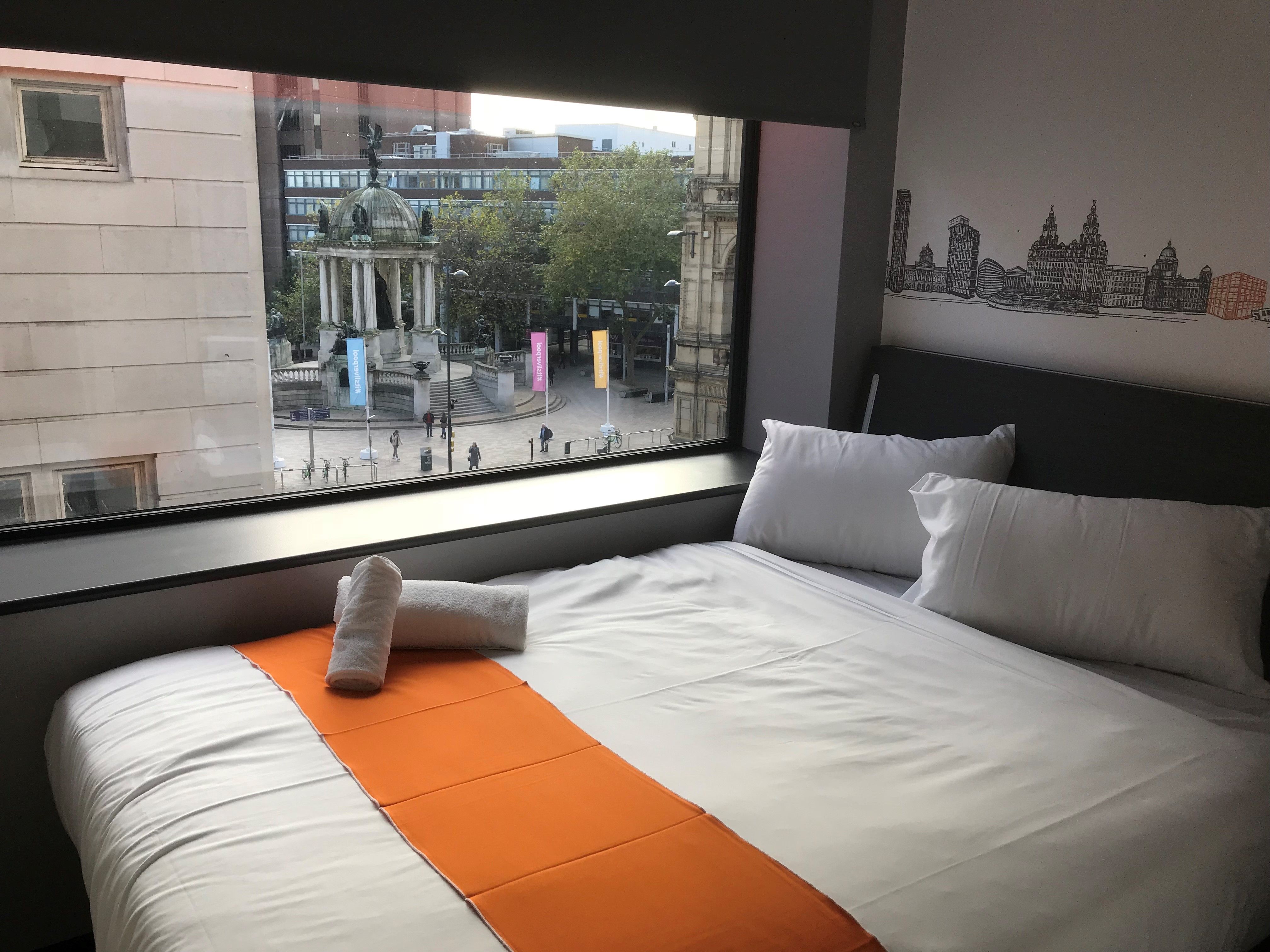 An easyHotel room in Liverpool with a white bed, orange runner, and a wall mural of the city skyline, overlooking the Queen Victoria Monument in Derby Square.