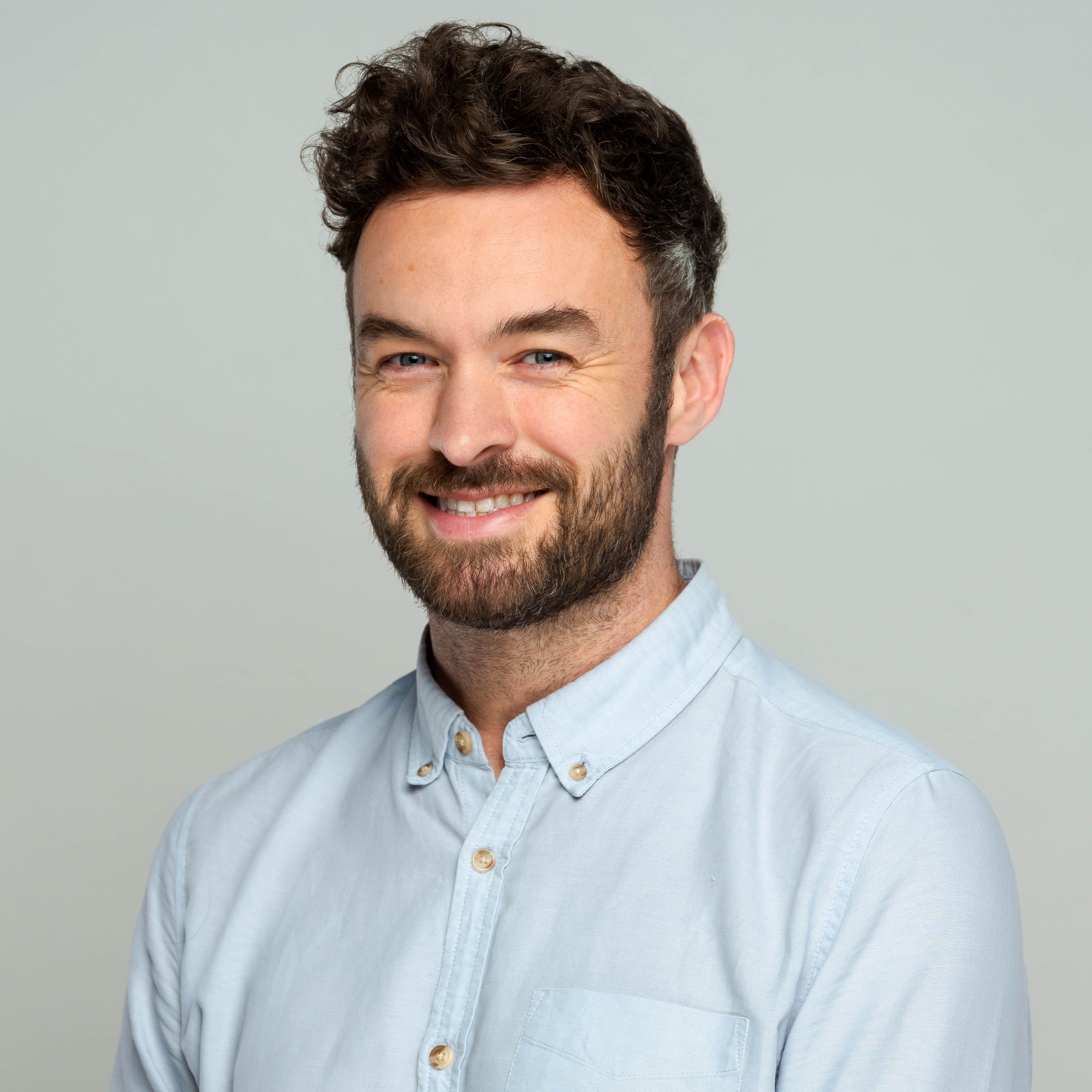 Professional headshot of Daniel Thompson, a smiling man with dark curly hair and a beard, wearing a light blue collared shirt.
