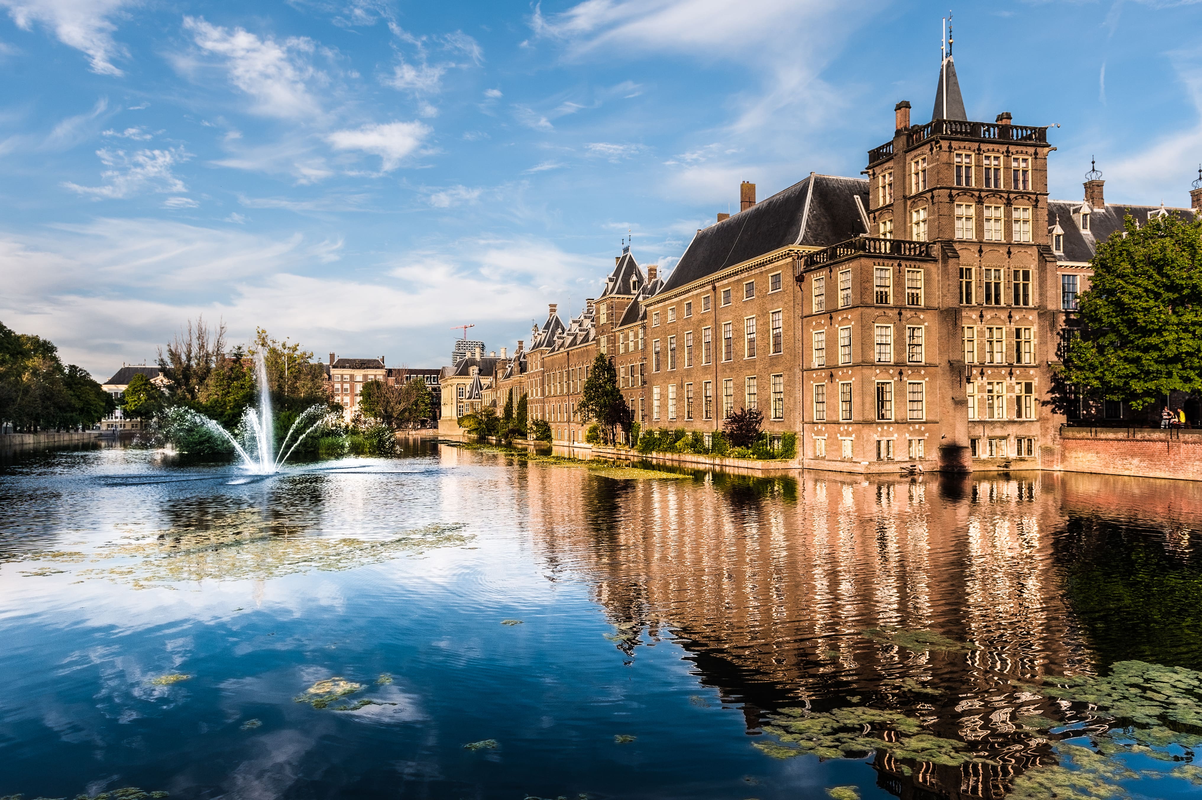 Historic Binnenhof parliament buildings in The Hague, Netherlands, reflected in the Hofvijver pond with a prominent fountain under a bright blue sky.
