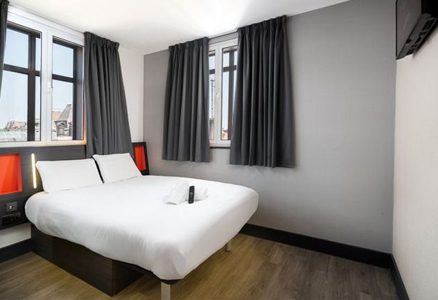 easyHotel room with a double bed, white duvet, two large windows with grey curtains, light grey walls, and wood-effect flooring. Features include modern bedside lighting, orange headboard accents, and a wall-mounted TV.
