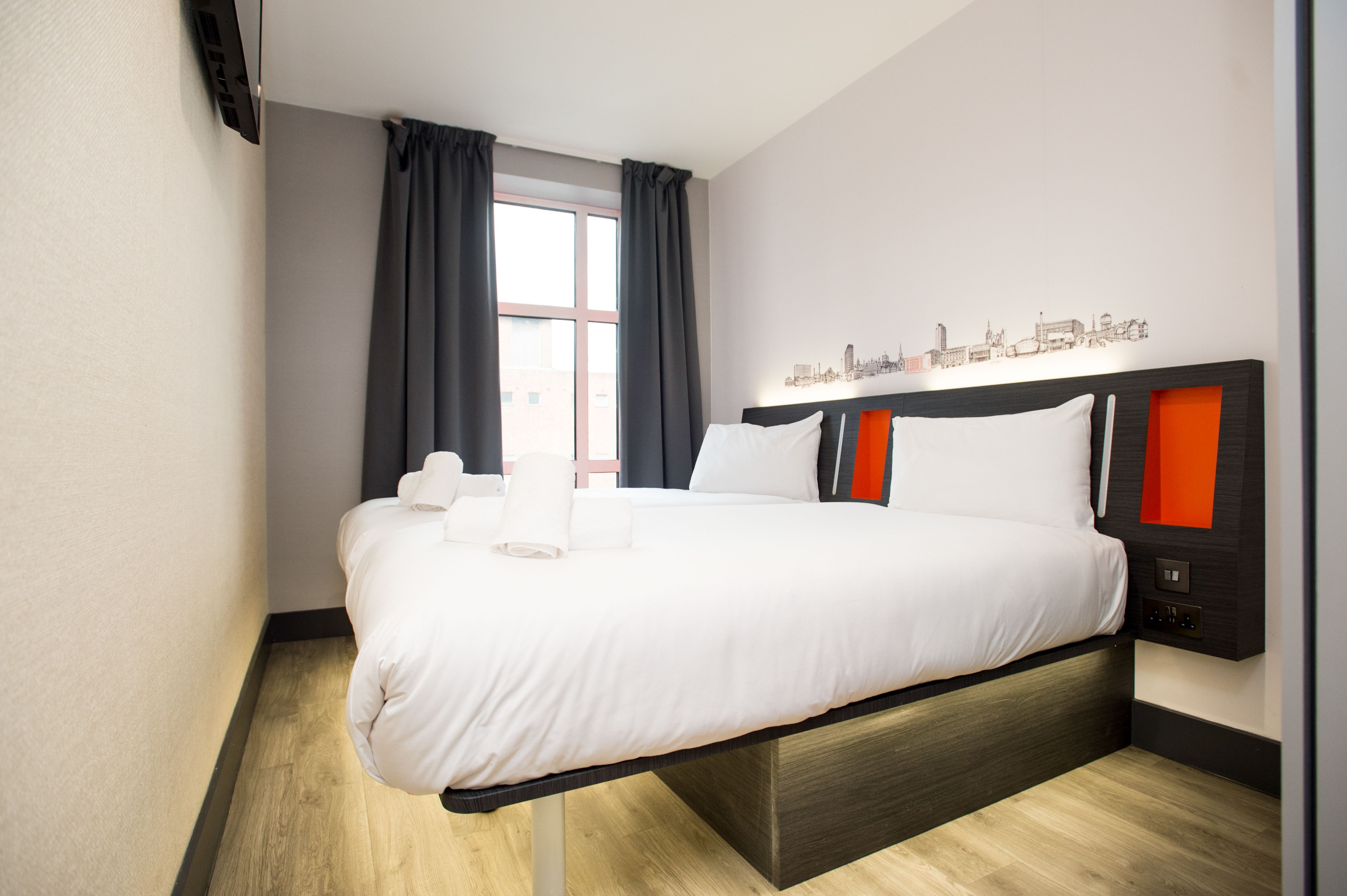 A modern, compact easyHotel room in London featuring a comfortable double bed with crisp white linens, a dark headboard with vibrant orange alcoves, and a unique London cityscape mural above. The room has light wood-effect flooring and dark grey curtains.