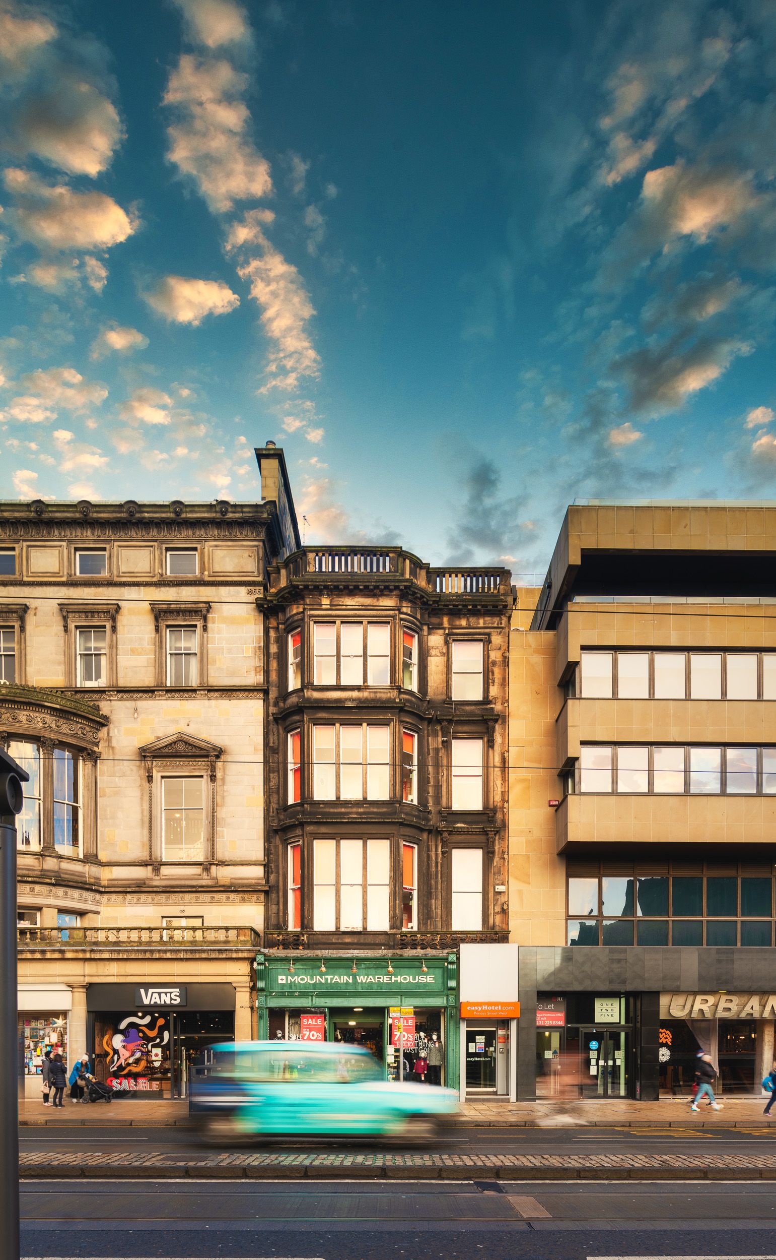 Edinburgh city street view featuring a mix of historic stone architecture and modern buildings, including a visible easyHotel.com entrance, Mountain Warehouse, and Vans stores. A blurred vehicle moves on the street under a dynamic blue sky with clouds.