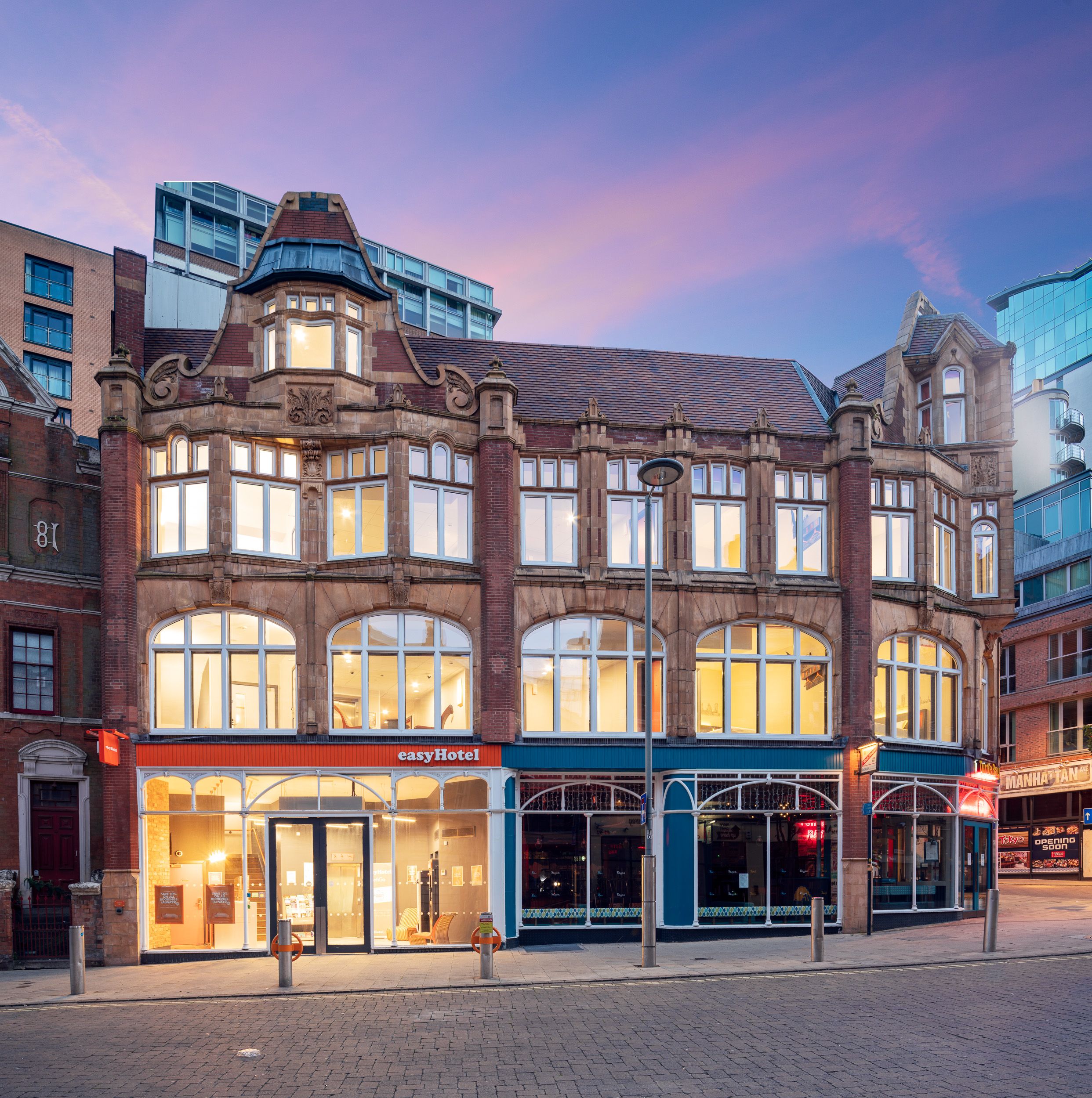 easyHotel Manchester building exterior on Dale Street at dusk, showcasing an ornate Victorian-style brick facade with large, lit windows under a vibrant pink and purple sky.