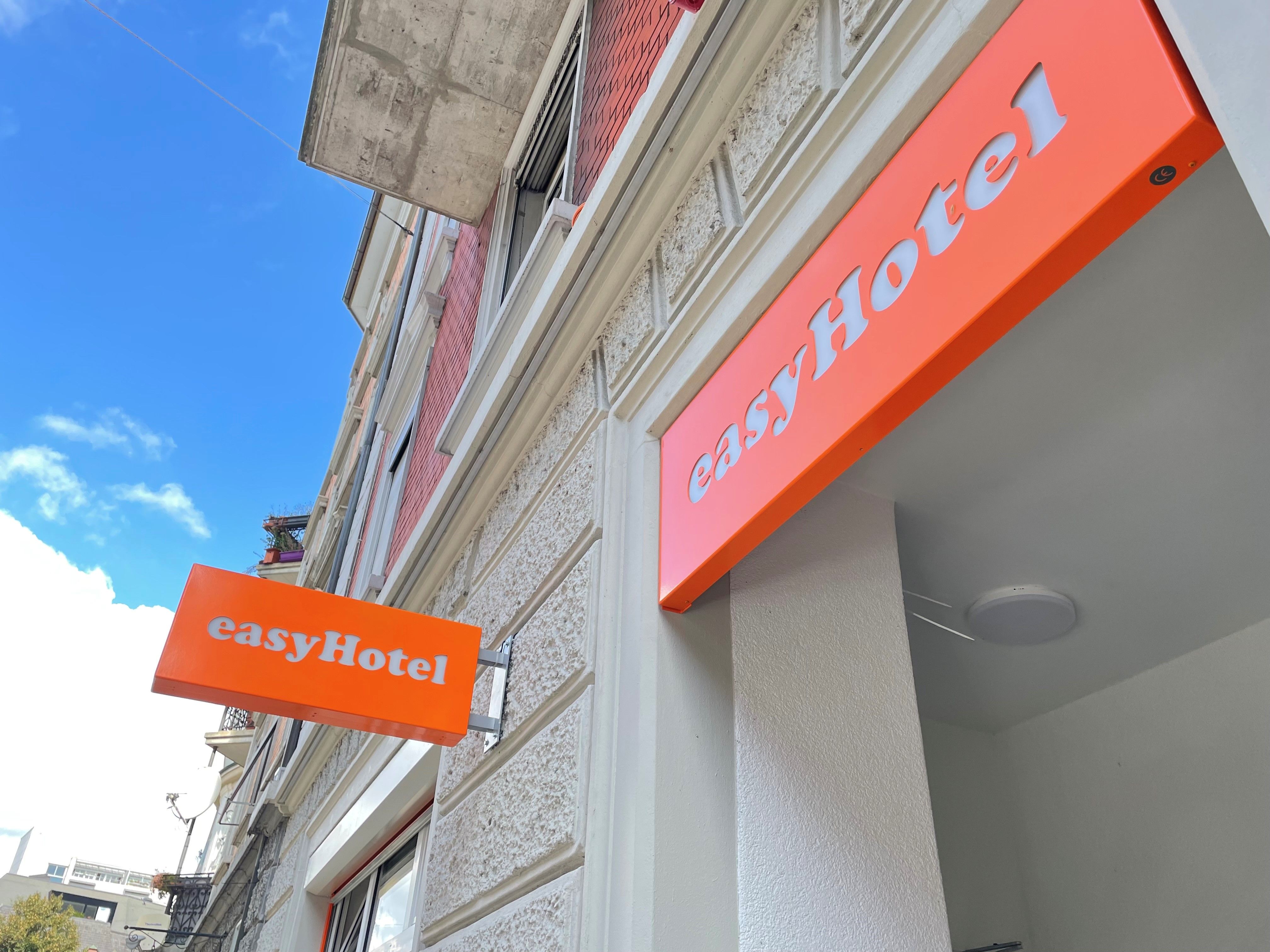 easyHotel building exterior with two prominent orange signs on a facade featuring light classical architectural details and red brickwork under a blue sky.
