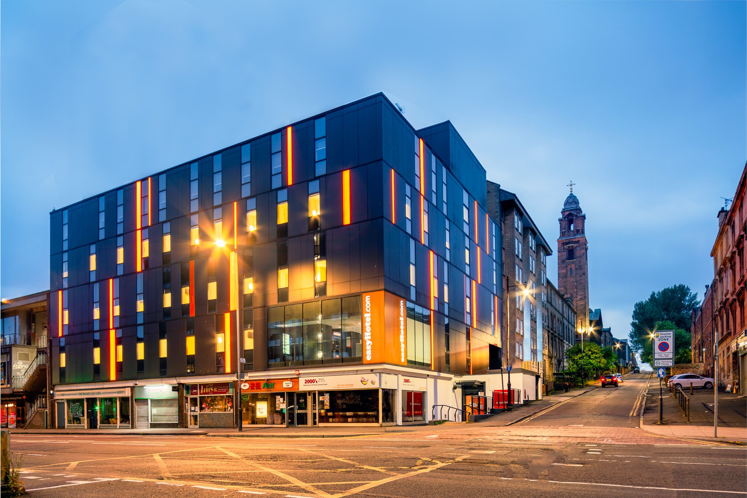 easyHotel Glasgow exterior at dusk, a modern building with dark paneling and vertical orange light strips, set on a city street with a historic stone tower in the background.