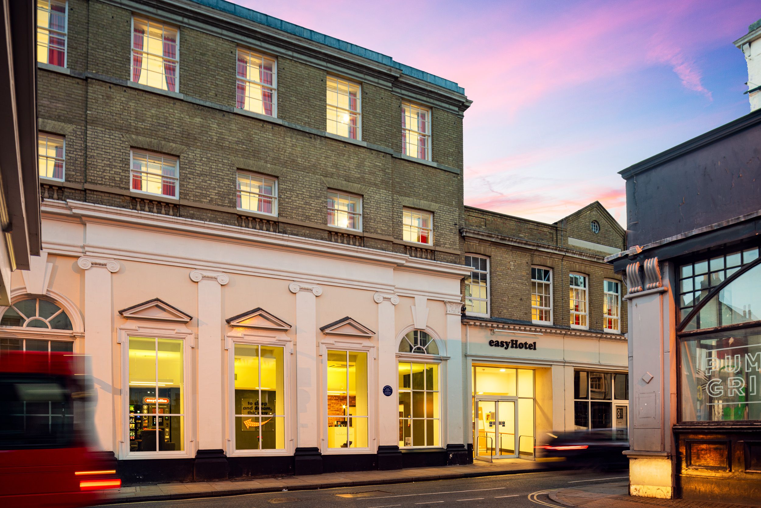 Exterior of easyHotel London Victoria at dusk, showing its brick and cream facade with illuminated windows. A red double-decker bus is visible on the street, against a vibrant pink and purple twilight sky.