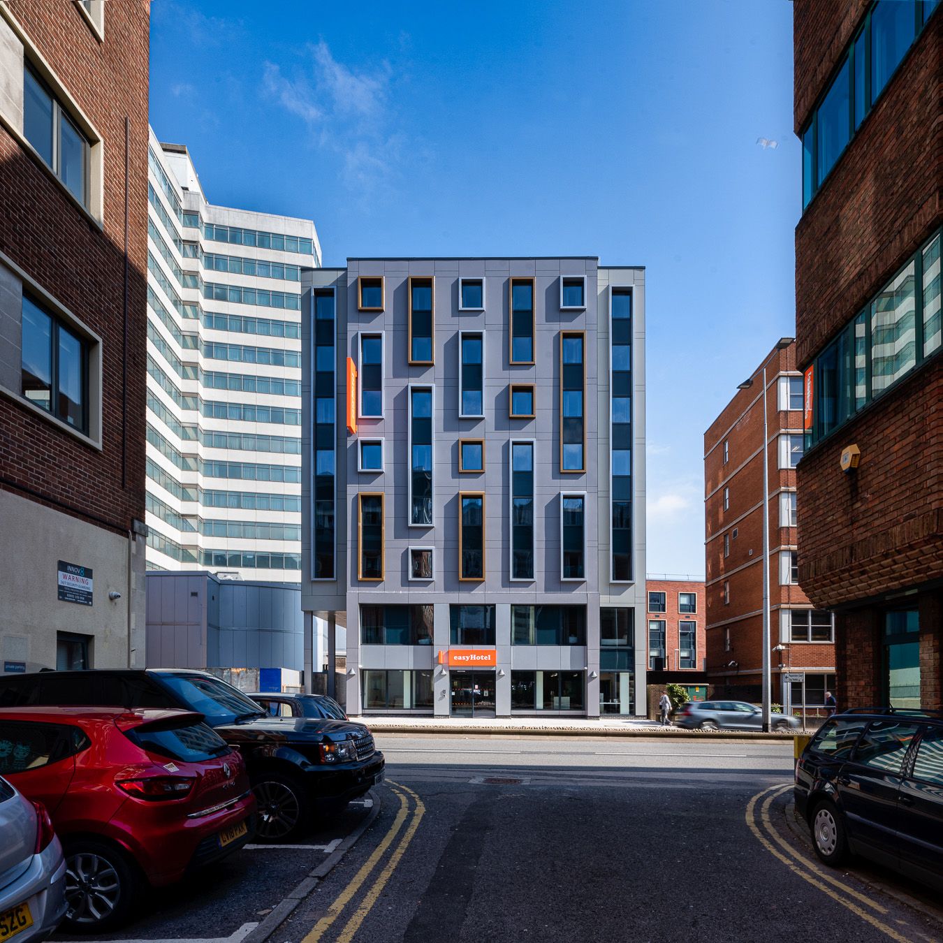 Exterior view of the modern easyHotel Southampton building from a city street, featuring a grey facade with numerous rectangular windows and the orange easyHotel logo. Parked cars are visible along the roadside, with other urban buildings under a clear bl