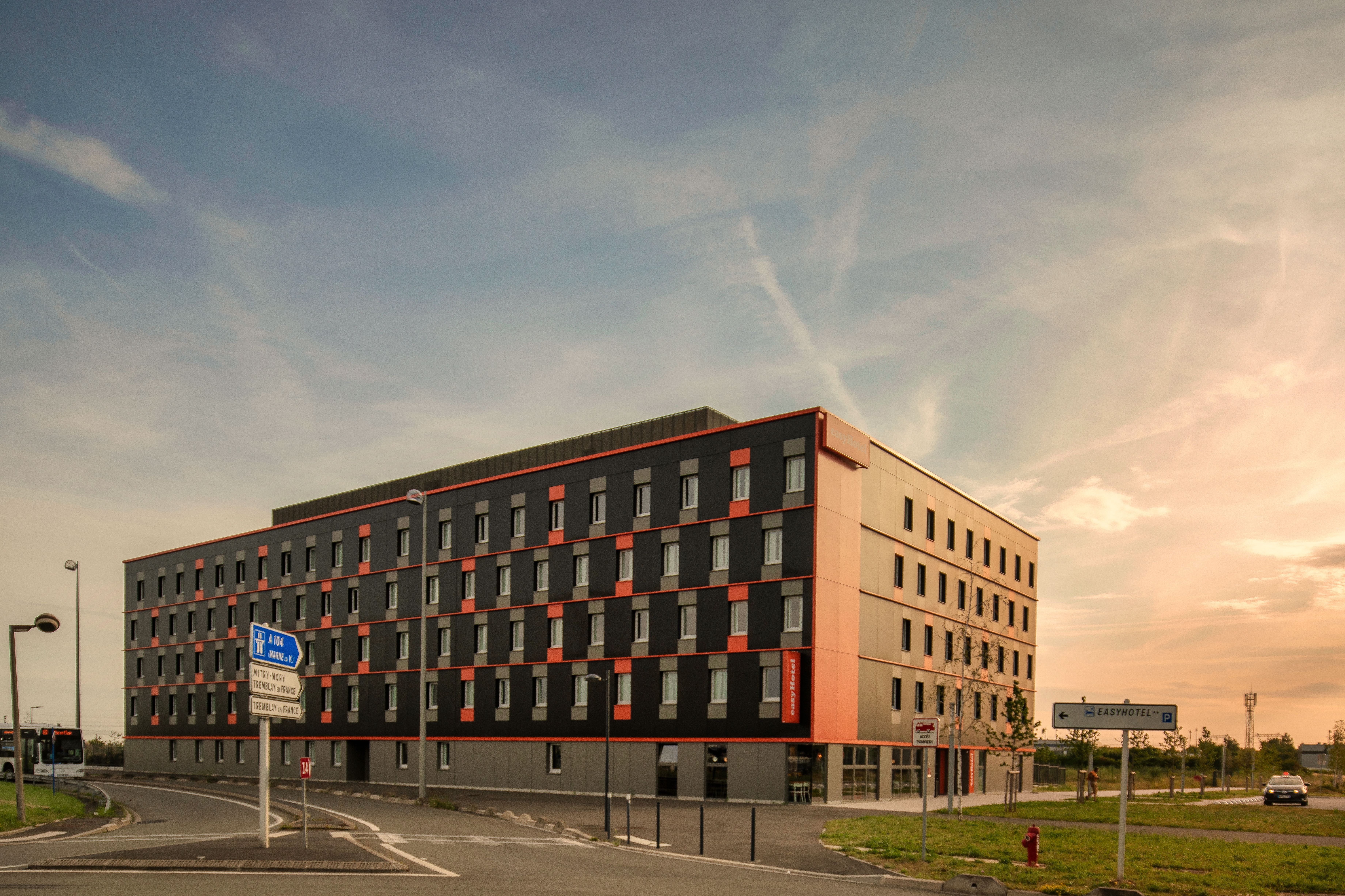 Modern easyHotel building exterior with a distinctive dark grey and orange facade under a sunset sky, located near Paris Charles de Gaulle Airport.