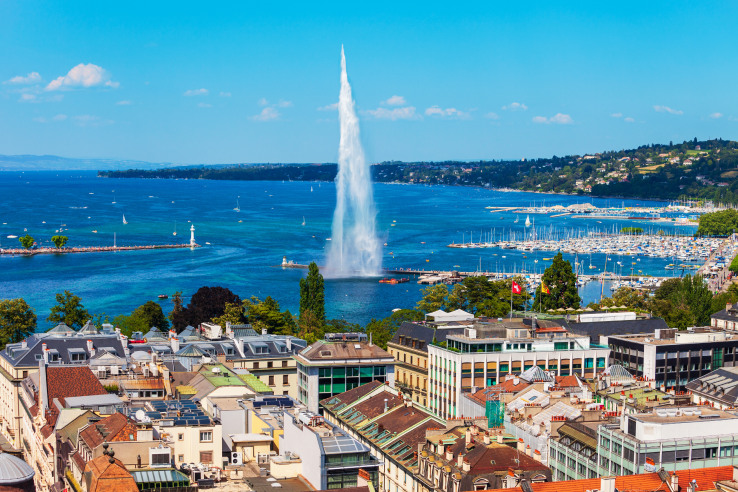 Panoramic view of Geneva, Switzerland, showcasing the iconic Jet d'Eau fountain on Lake Geneva, a lively marina with sailboats, and the vibrant city skyline under a blue sky.