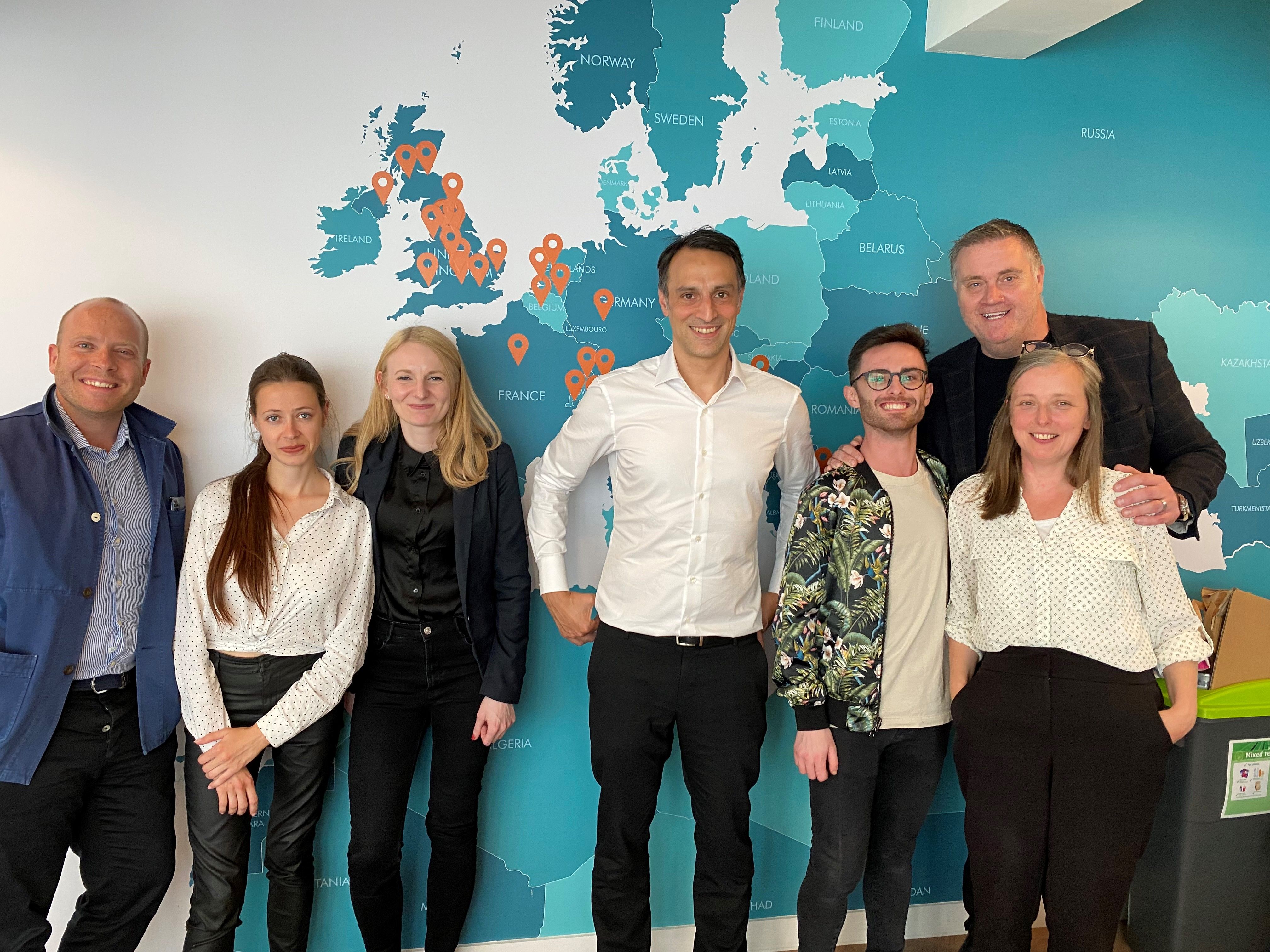 Group photo of seven smiling business professionals standing in an office with a large map of Europe on the wall, featuring orange pins indicating multiple locations across the UK and mainland Europe.
