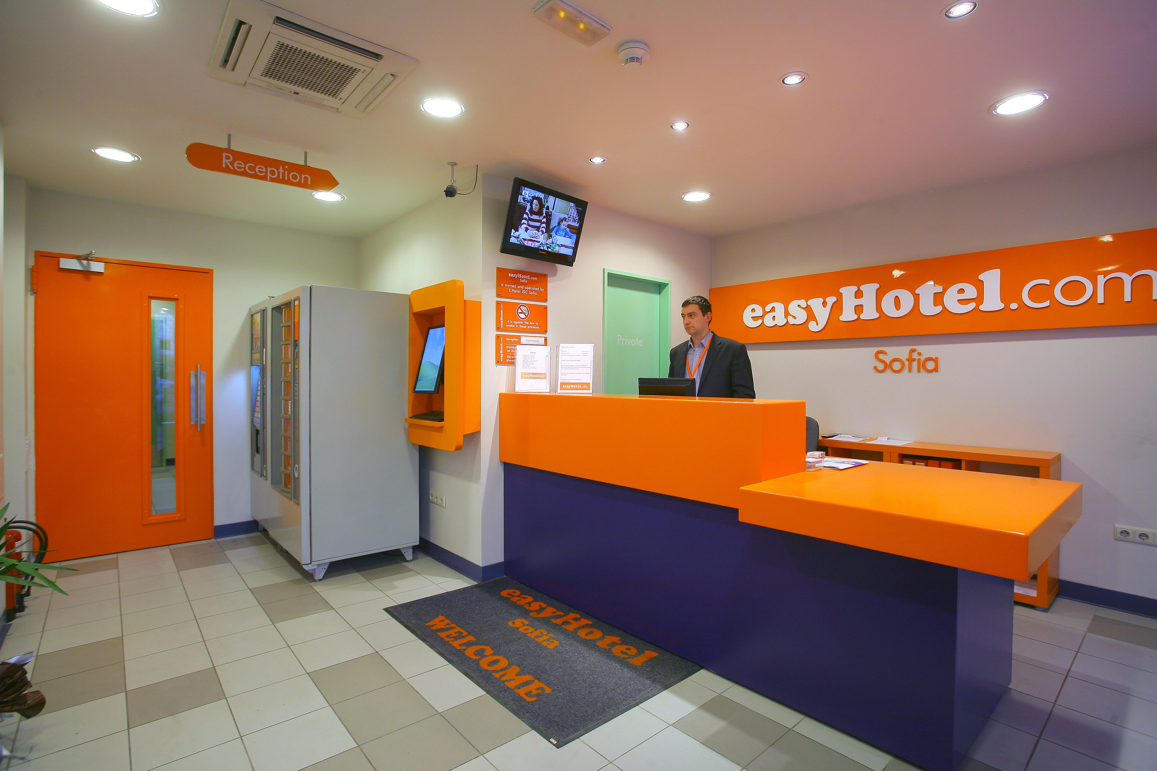 easyHotel Sofia reception area with a staff member behind an orange and blue desk, and visible easyHotel branding.