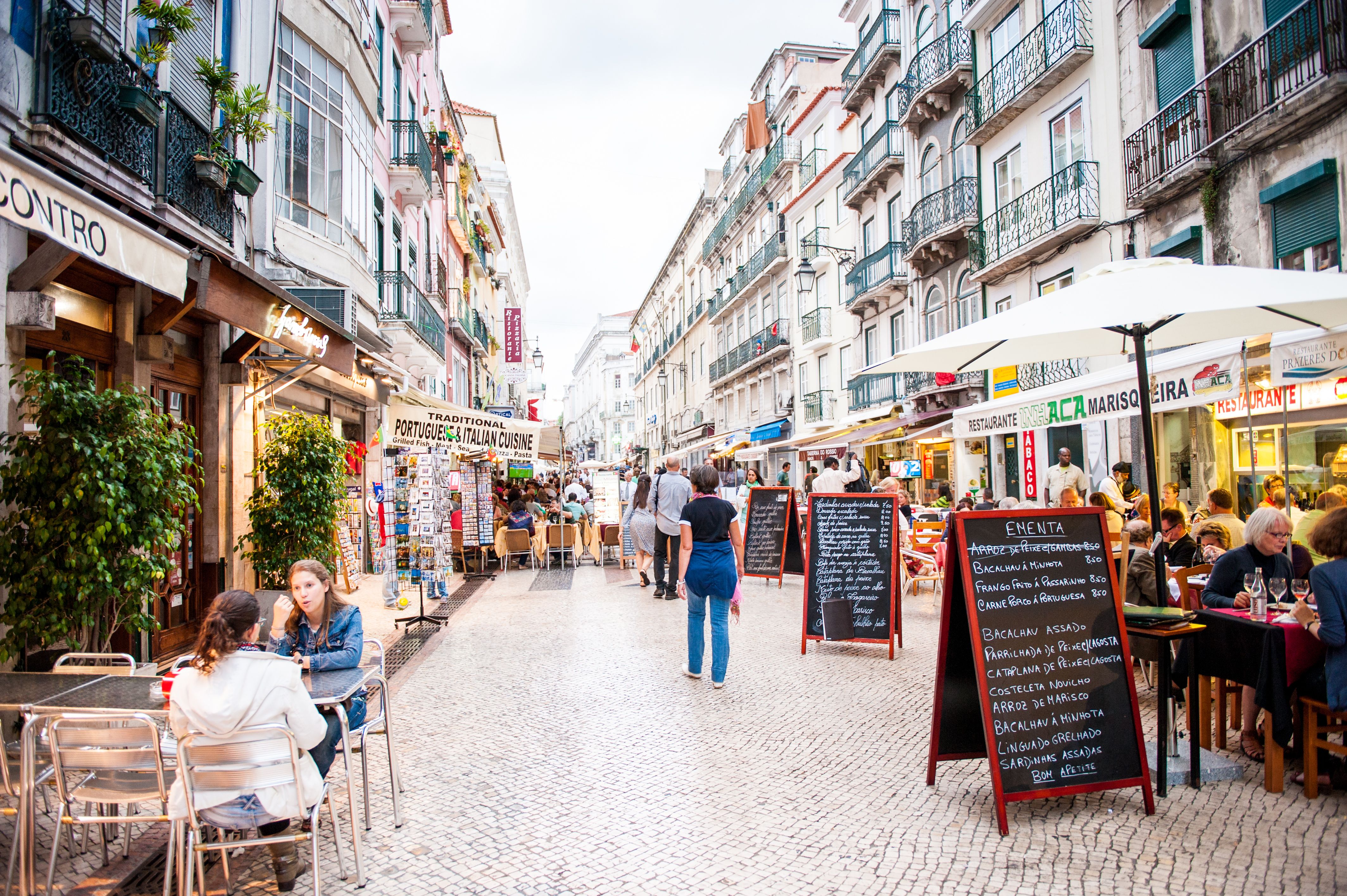 Bustling pedestrian street in Lisbon, Portugal, featuring numerous restaurants with outdoor dining tables and people enjoying meals. Historic buildings line the cobblestone street, with menu boards displaying Portuguese cuisine.