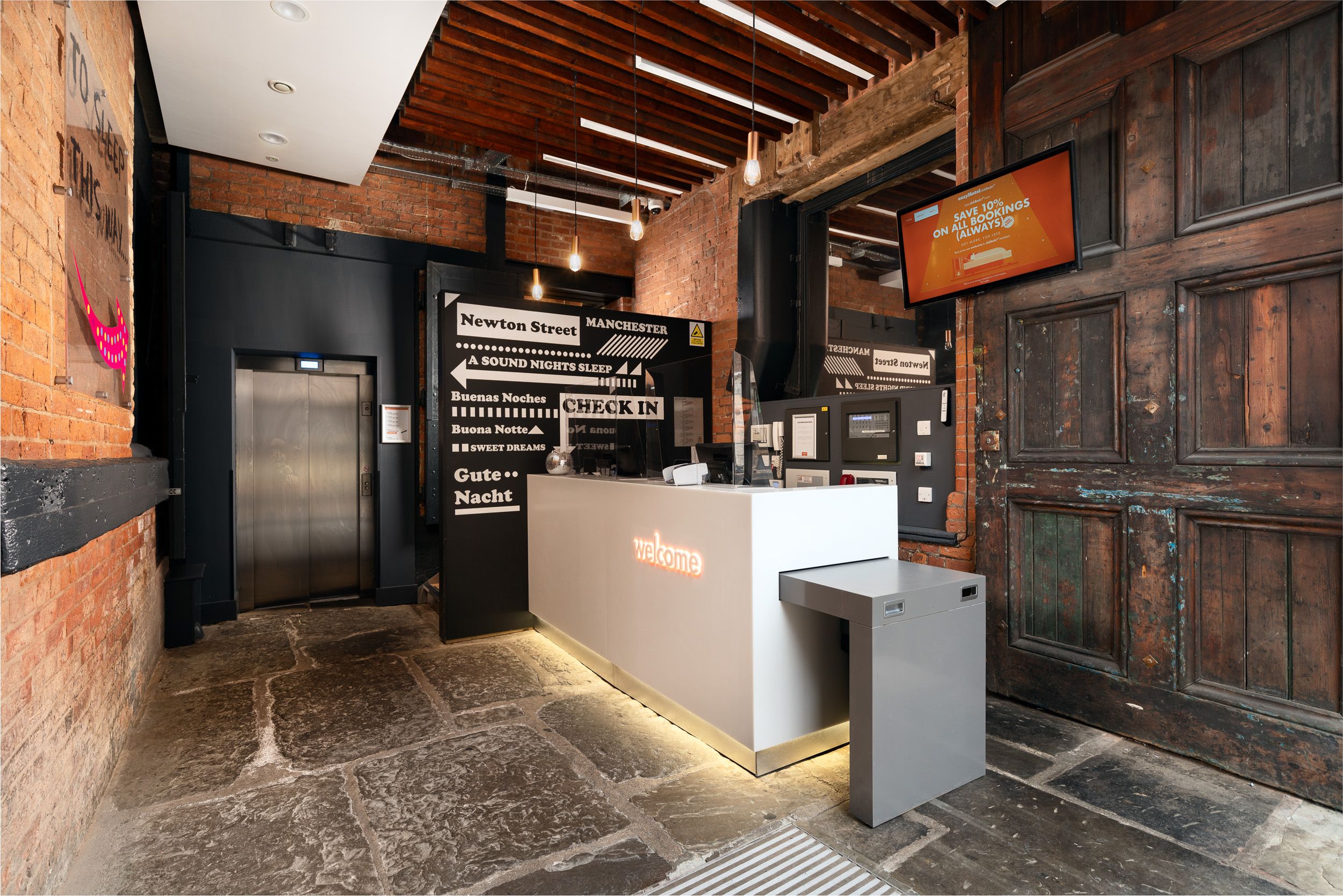 easyHotel Manchester Newton Street lobby with a modern self-service check-in desk, industrial exposed brick walls, stone floor, and an elevator.