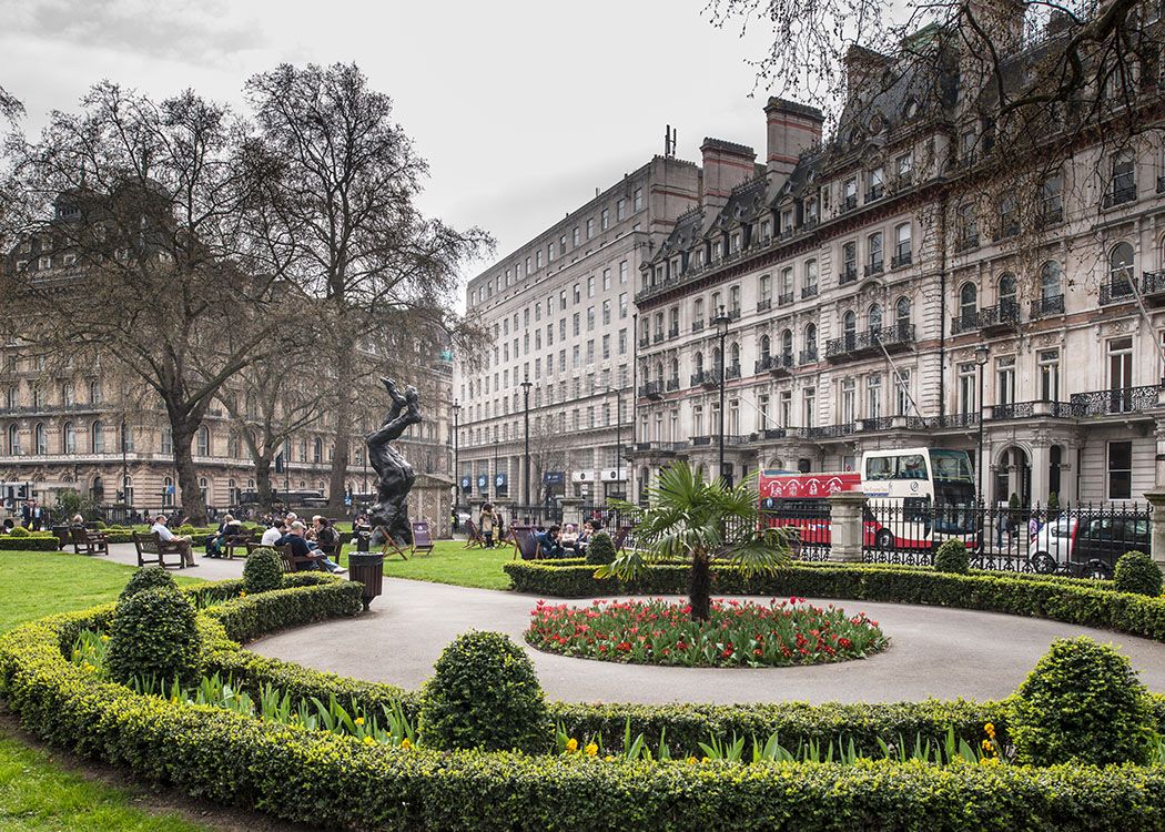 Grosvenor Gardens in London, featuring the bronze 'The Dance' sculpture, a red double-decker bus, and ornate historic buildings surrounding a public park with people relaxing on benches.