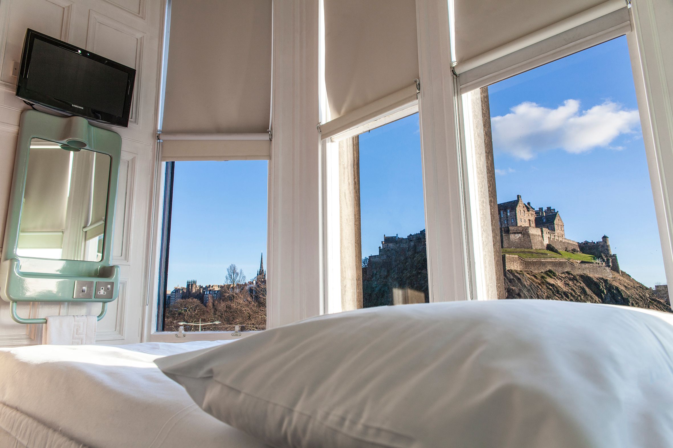 Panoramic view from a hotel room in Edinburgh, Scotland, featuring a bed, TV, and a bright interior looking out to the majestic Edinburgh Castle on a sunny day.