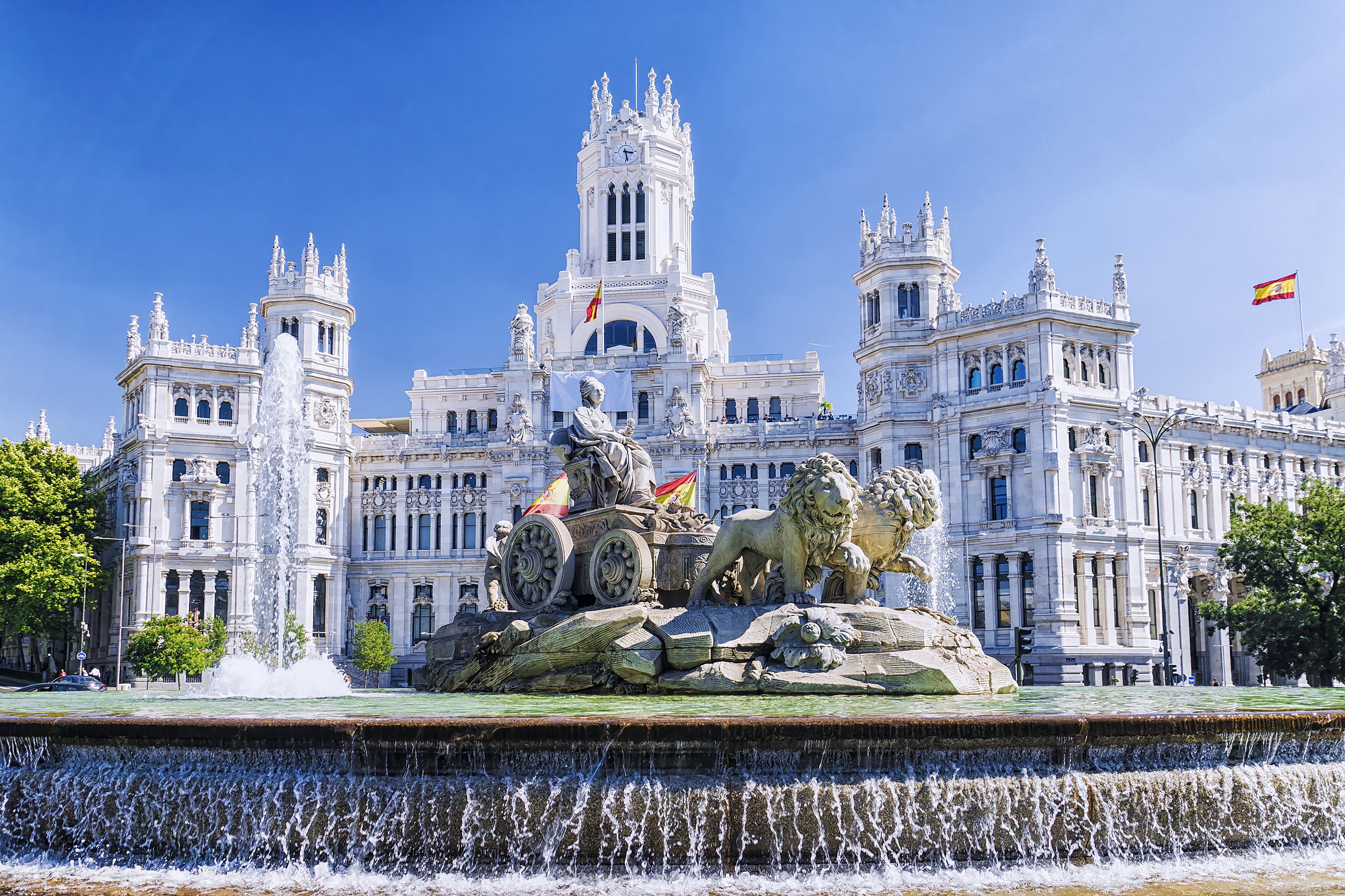 The iconic Cibeles Fountain with statues of Cybele and lions, set against the grand Cybele Palace (Madrid City Hall) under a clear blue sky in Madrid, Spain.
