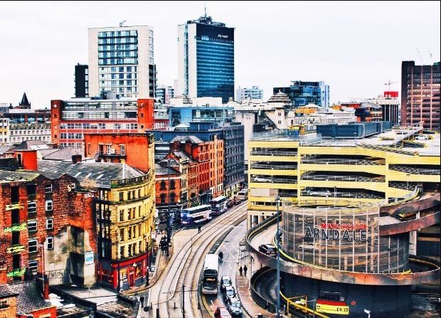 Elevated view of Manchester city centre, featuring tram lines winding through a mix of historic and modern buildings, with the distinct circular Manchester Arndale car park prominent in the foreground.