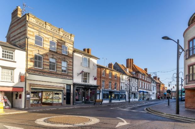 Historic high street in the Milton Keynes area, featuring a row of traditional brick and timber-framed buildings housing shops under a clear blue sky, with a small roundabout in the foreground.