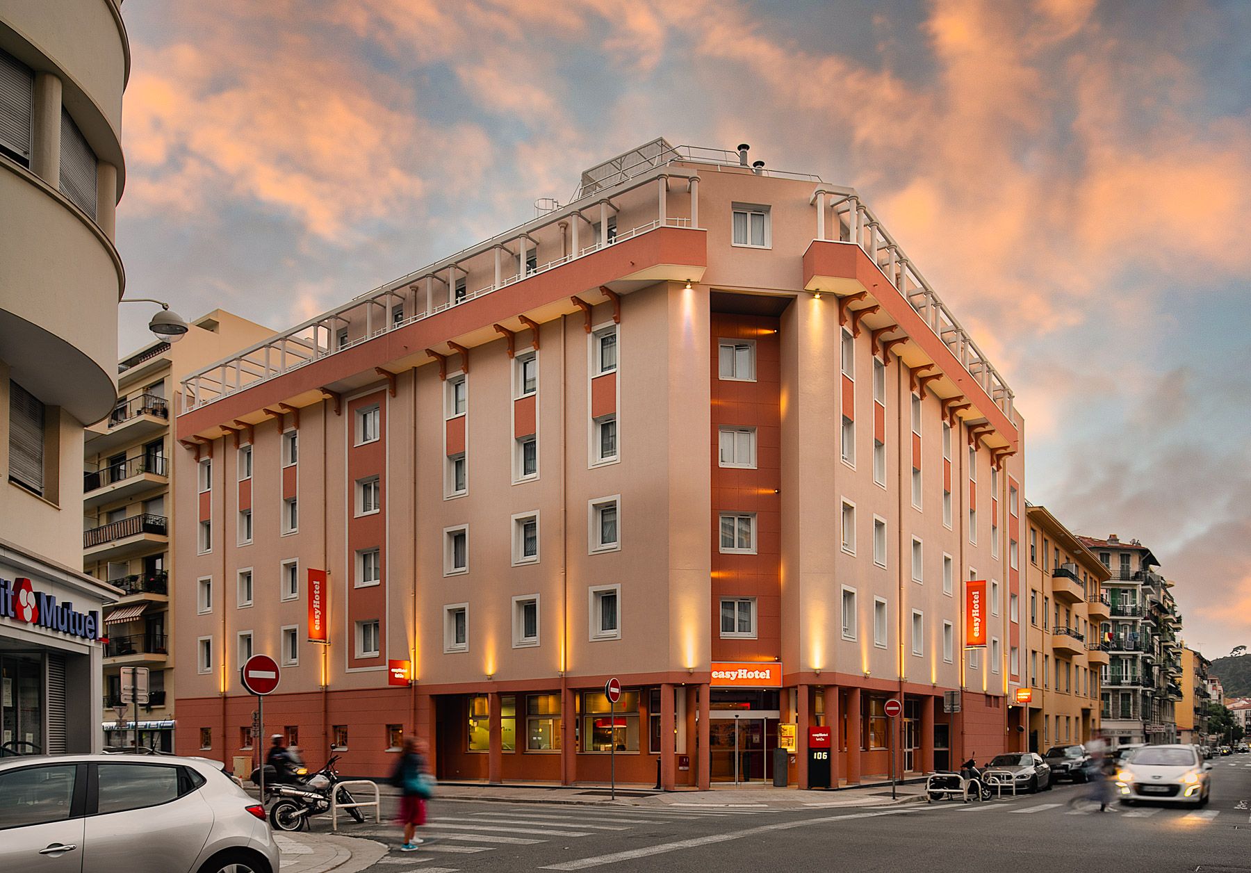 Exterior view of the easyHotel in Nice, France, featuring its peach and orange facade at dusk. The multi-story building stands prominently on a street corner, with warm lights illuminating the entrance and surrounding urban street scene.