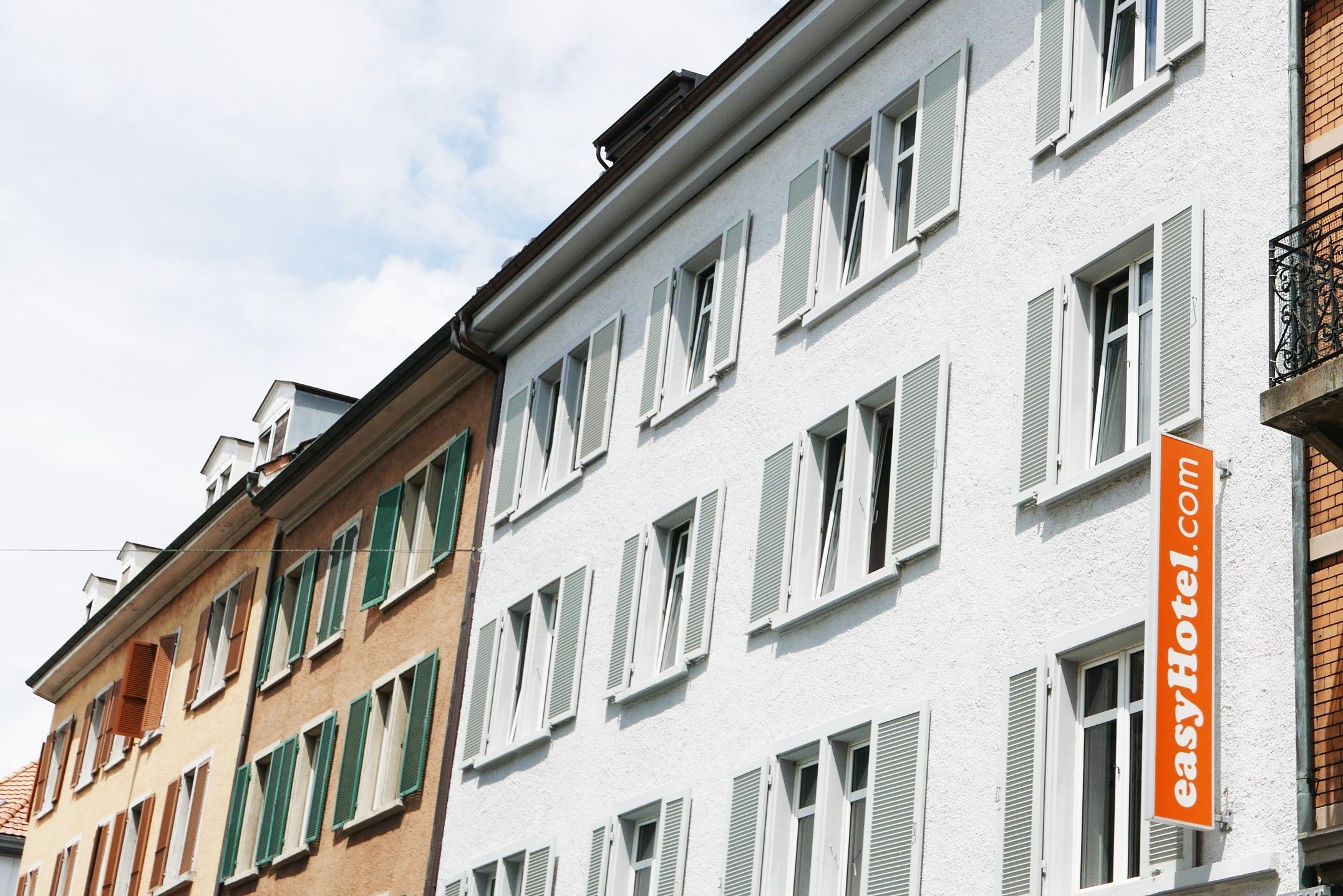 EasyHotel building exterior in a European city, featuring white stucco walls, windows with light grey shutters, and an orange easyHotel.com vertical banner.