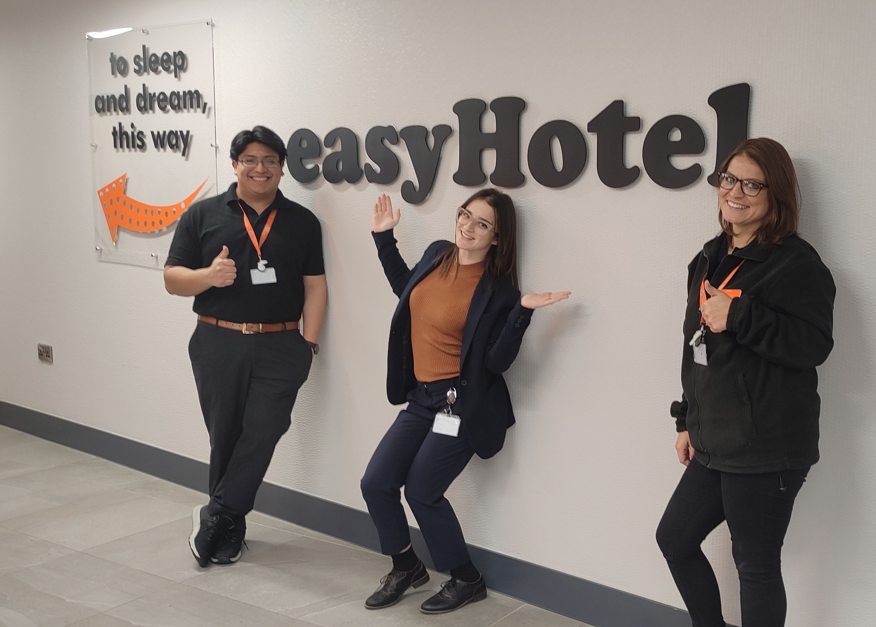 Three smiling easyHotel staff members in the lobby of easyHotel Oxford, posing in front of the easyHotel logo and a sign reading 'to sleep and dream, this way'.