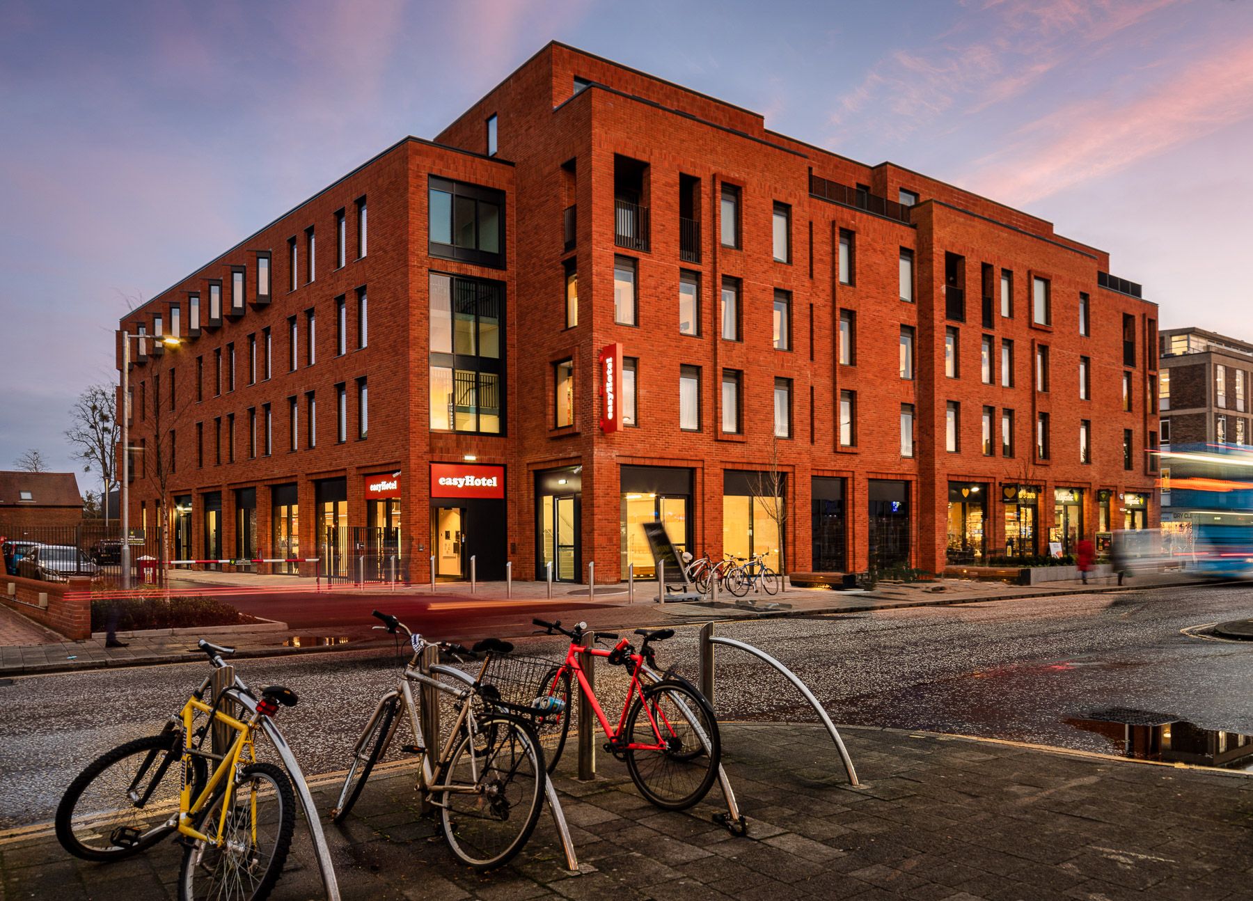 EasyHotel Oxford building exterior at dusk, a modern red brick hotel with large windows, featuring parked bicycles and a passing blurred bus on the street.