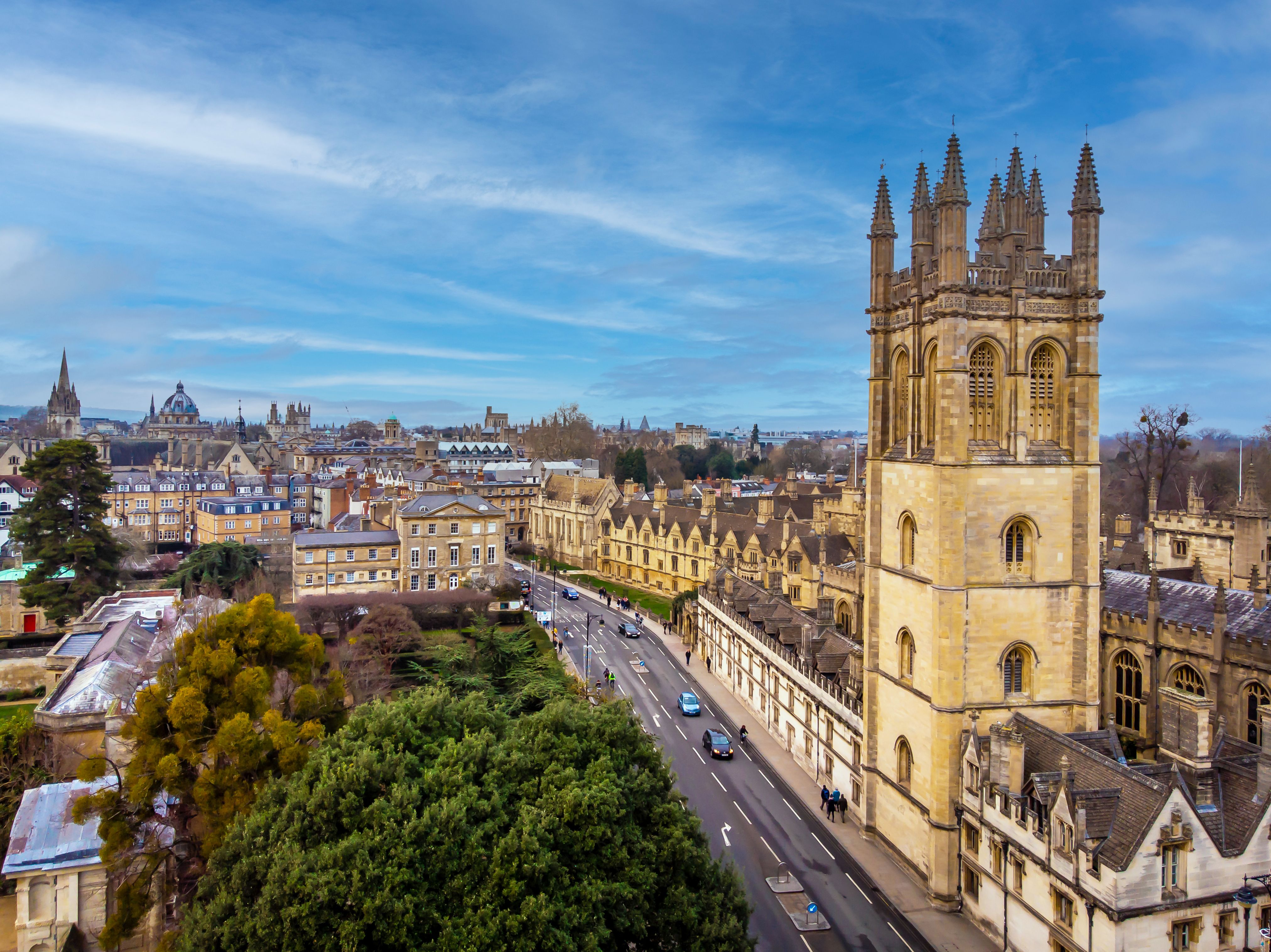 Aerial view of Oxford city skyline featuring iconic university buildings including Magdalen College Tower, historic colleges, a street with cars and pedestrians, and green trees under a partly cloudy sky.