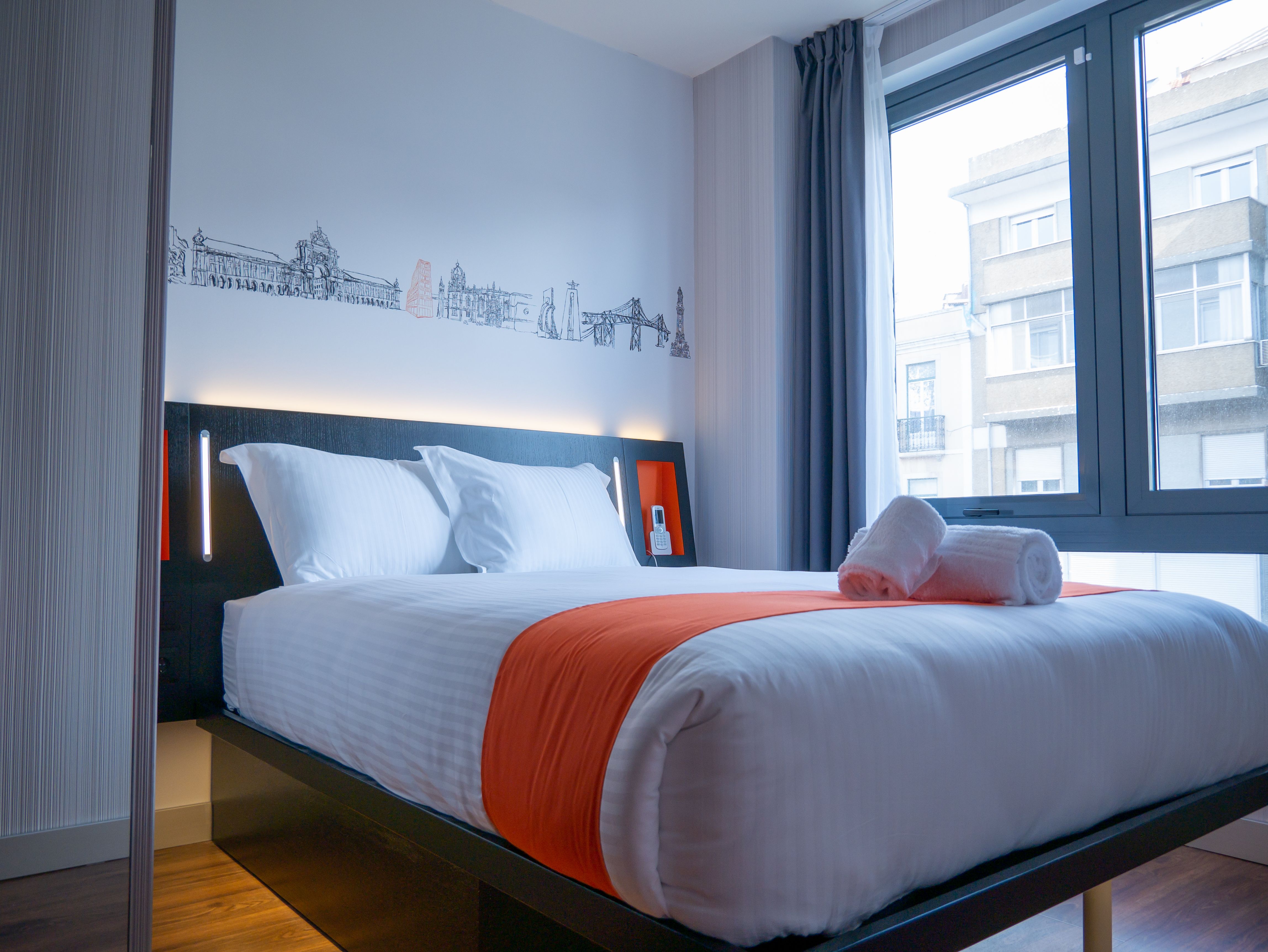 A modern easyHotel Porto room featuring a comfortable bed with white linens and an orange bed runner, alongside two rolled towels. A black line art mural depicting iconic Porto city landmarks decorates the white wall above the dark headboard. The room inc