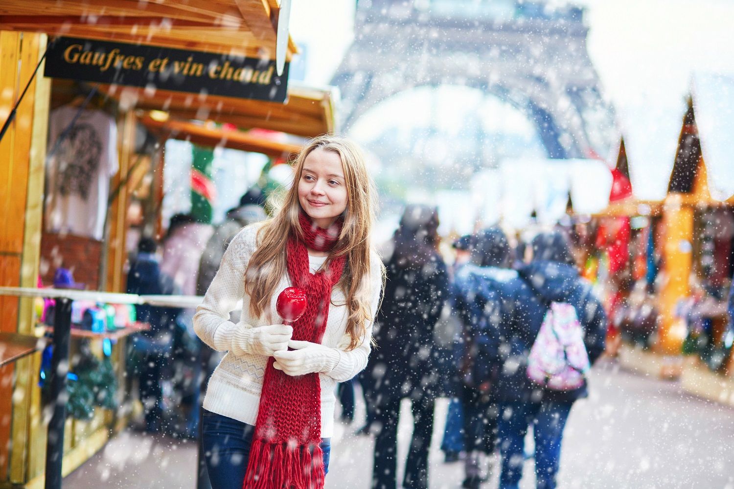 Smiling young woman holding a candy apple at a snowy Paris Christmas market, with the iconic Eiffel Tower visible in the background.