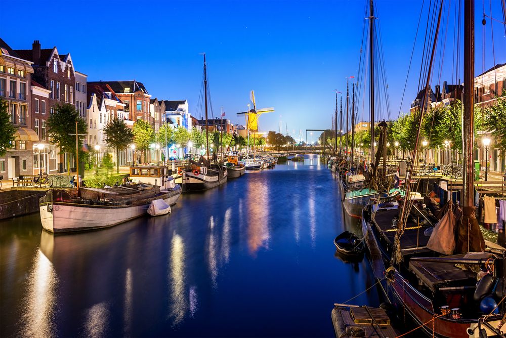 Historic Dutch canal in Delft at twilight, featuring traditional gabled buildings, numerous boats docked along the waterfront, and a prominent illuminated windmill reflecting on the dark water.