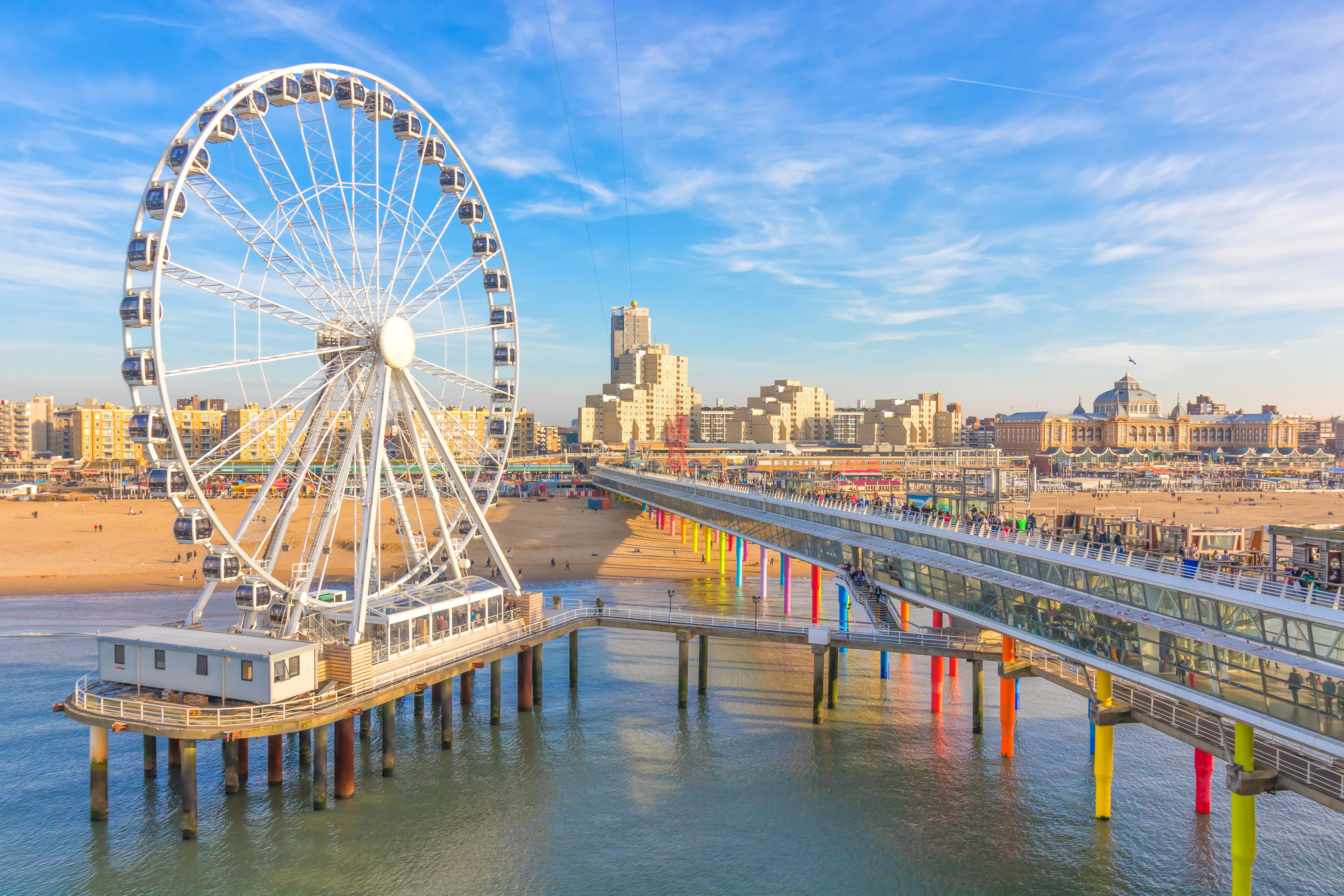 Scheveningen Beach pier with a large white Ferris wheel, colorful support columns, and the distant coastal skyline of The Hague under a blue sky.